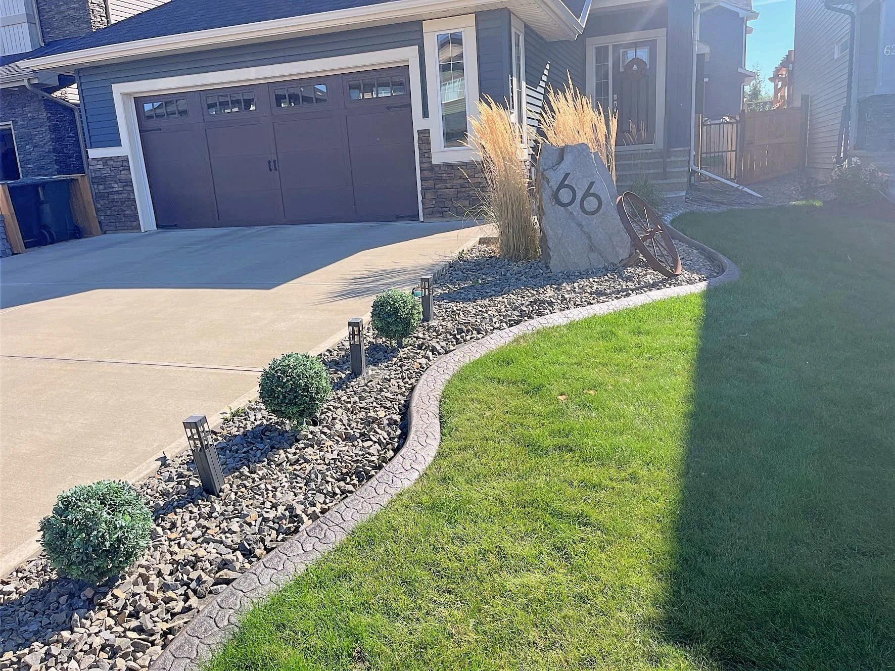 Front yard of a house with a curved lawn, a stone and gravel landscape bed with small bushes and pathway lights, a large stone with the number 66, and a bicycle leaning against the stone. The house has a blue exterior, a driveway, and a garage door.
