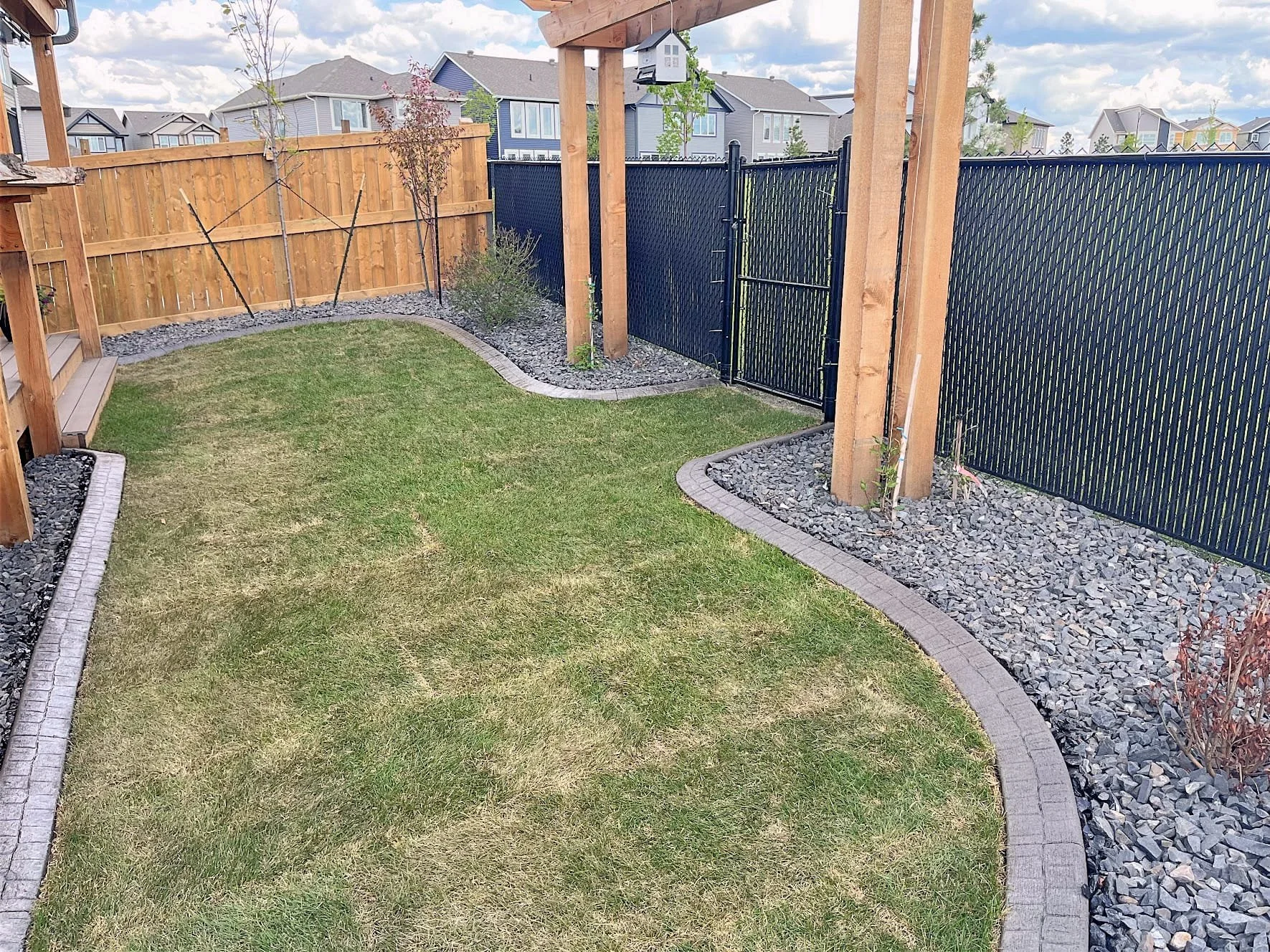 A backyard with a well-maintained lawn, bordered by a stone and brick edging, and a black metal fence with a gate. The yard features a wooden deck on the left side, a wooden fence in the background, some small trees, and a cloudy sky.