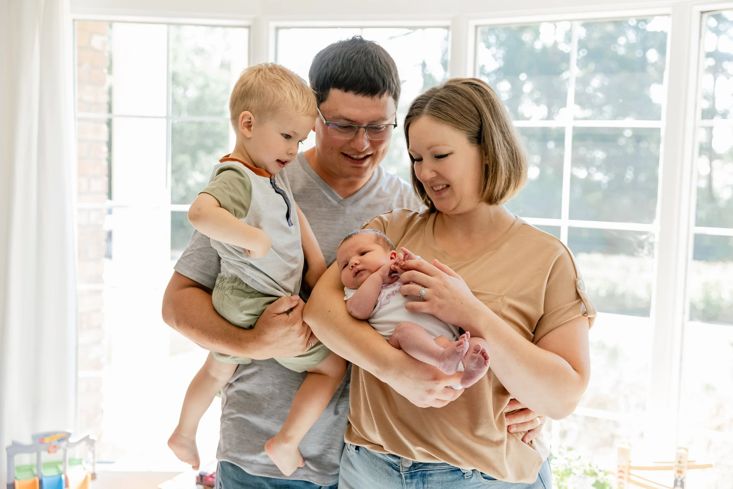 A family of four celebrating their newborn baby in a bright room with large windows, with the mother holding the baby and the father and older son looking at the baby affectionately.