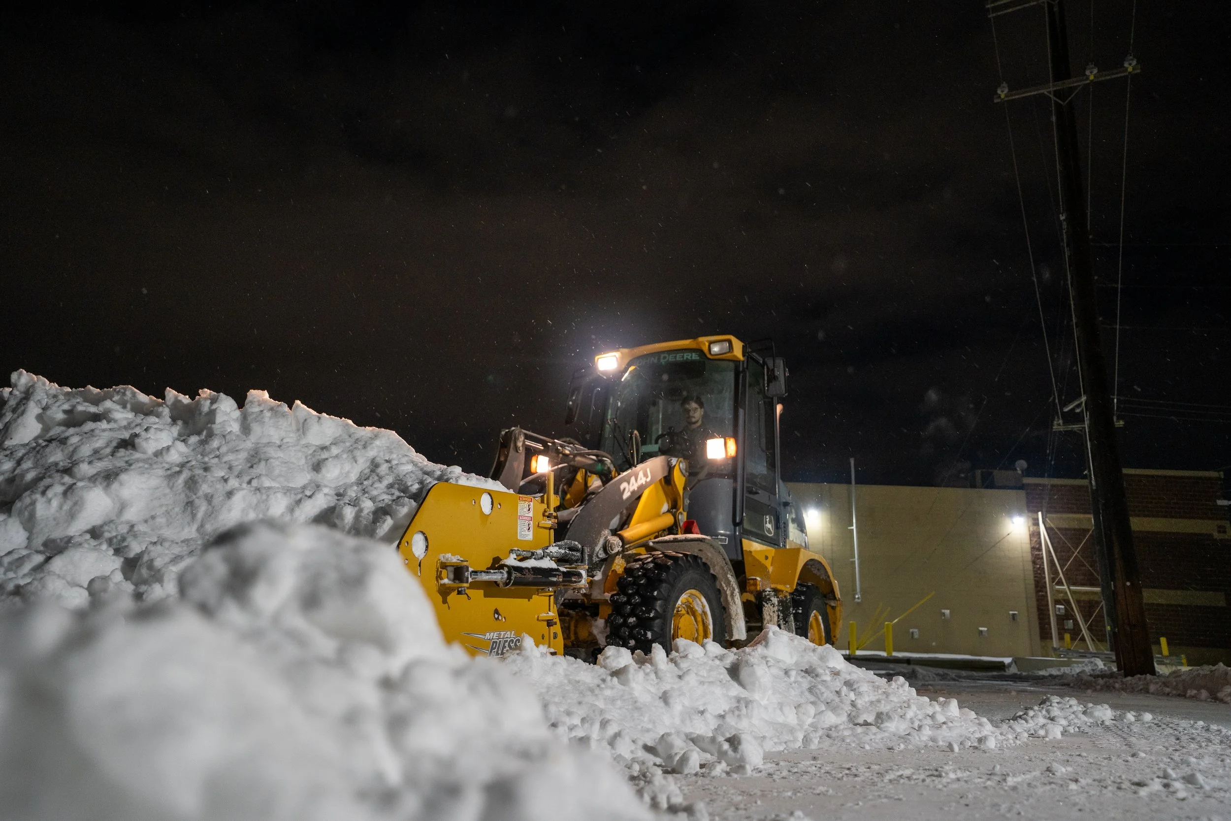A yellow John Deere snowplow clearing snow at night with a dark sky and a building in the background.