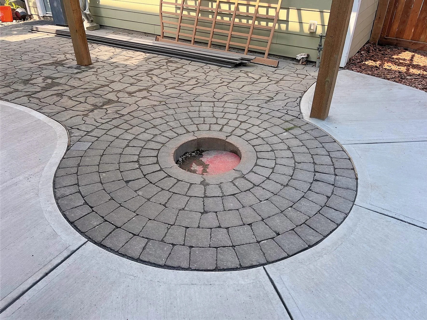 A circular stone patio with a small, empty fire pit in the center, surrounded by concrete paving slabs and a wooden post on the left side.