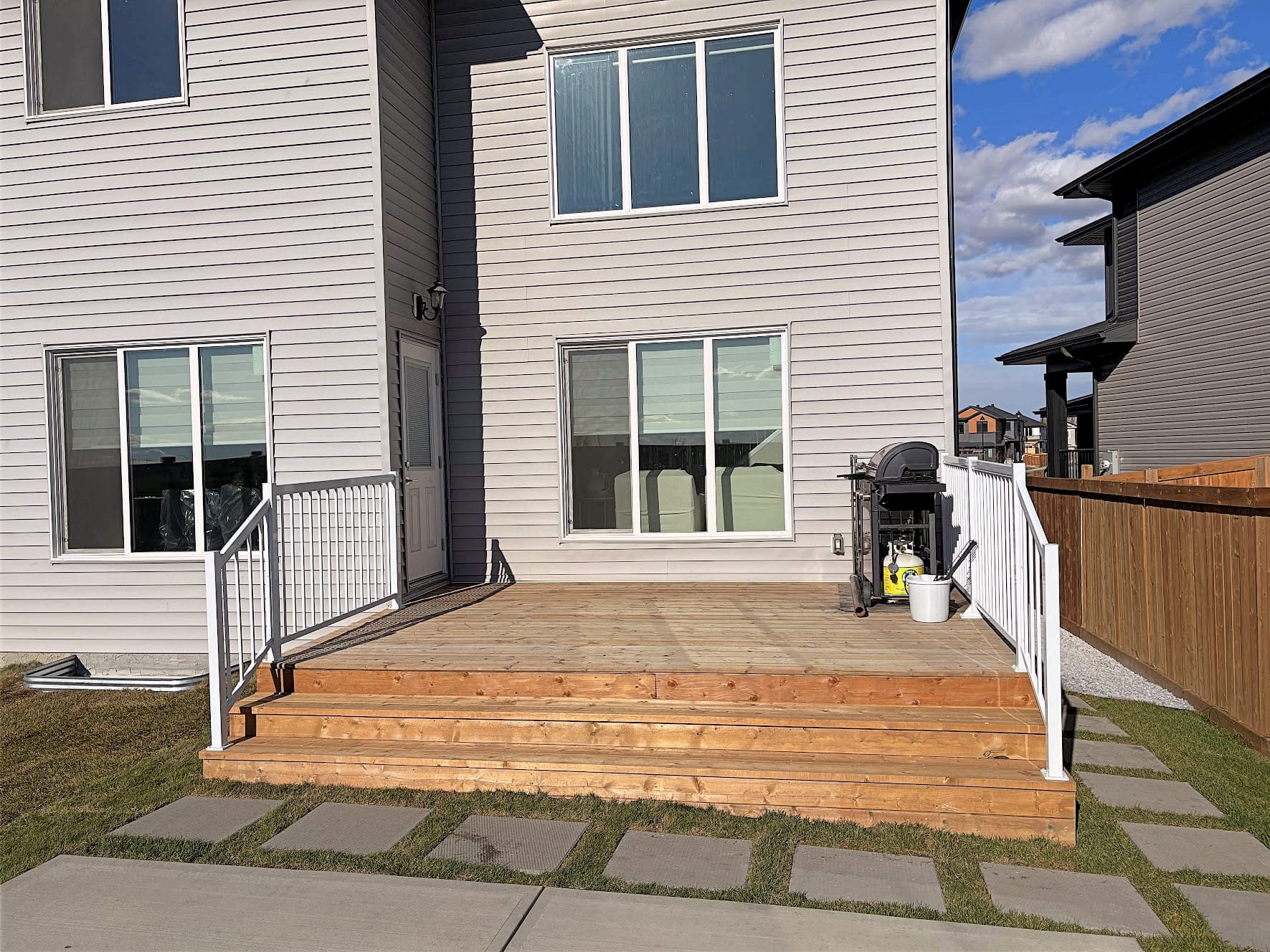 Backyard wooden deck with white railing, attached to a gray house with white trim, featuring steps leading to a concrete pathway, a gas grill with propane tank, and a white bucket, under a partly cloudy sky.
