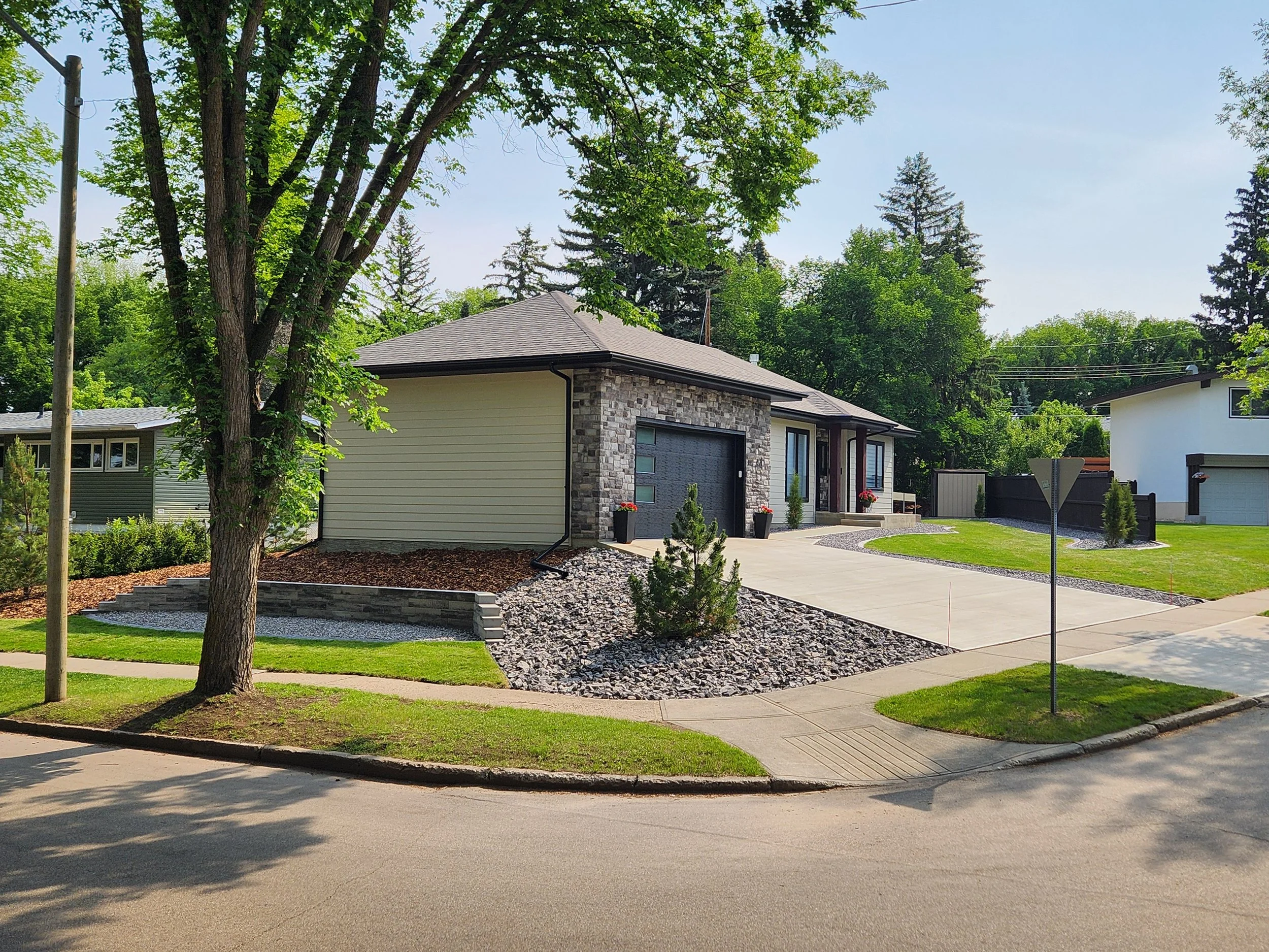 A modern single-story house with a stone and siding exterior, a gray garage door, and a concrete driveway. The front yard has landscaped rocks, small plants, and a well-maintained lawn, with a sidewalk and a tree in the foreground.
