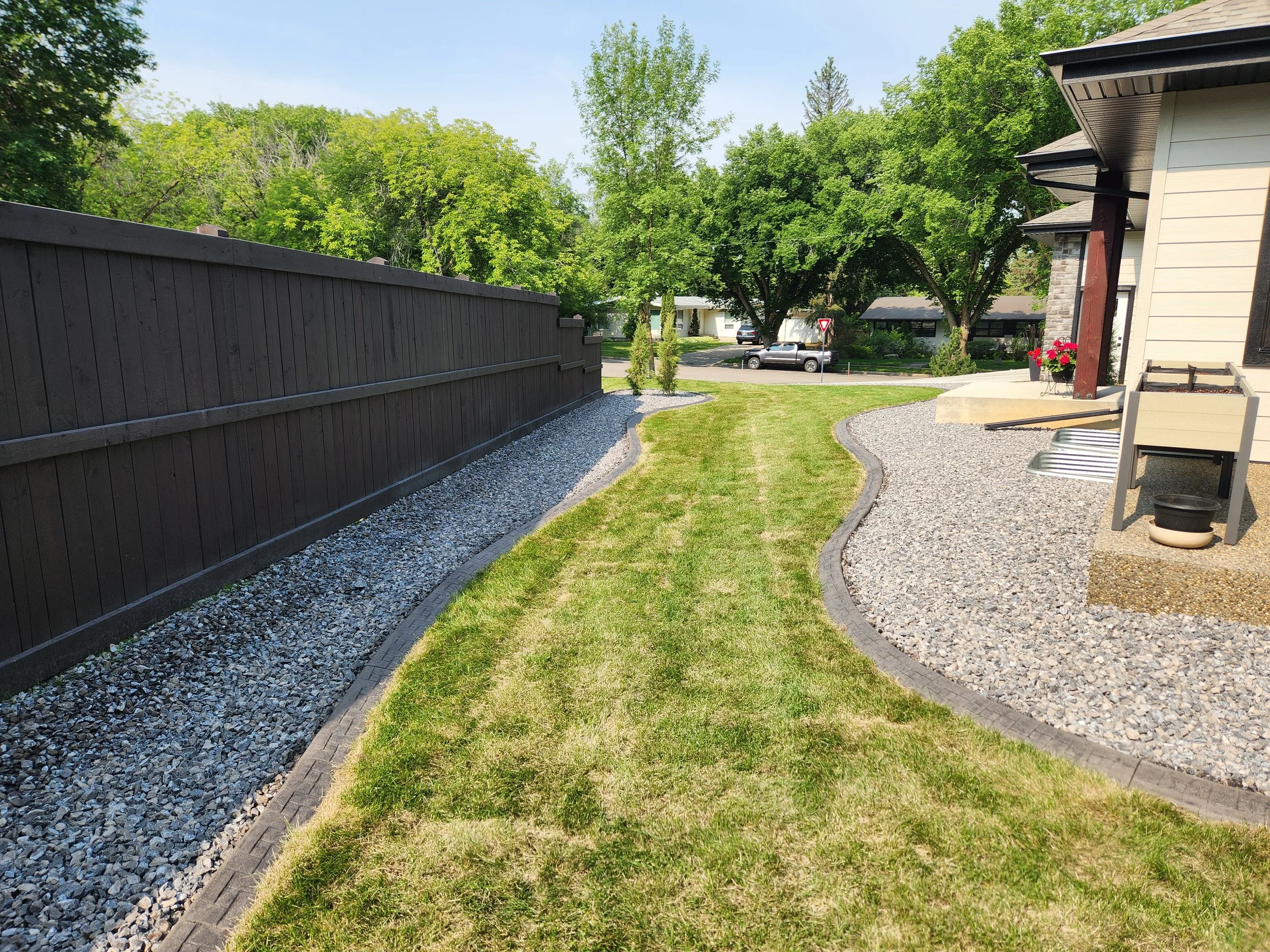 Backyard with a curved grassy pathway, bordered by decorative edging, surrounded by gravel areas, a dark wooden fence on the left, a house on the right with planters and a small patio area, and trees and houses across the street in the background.
