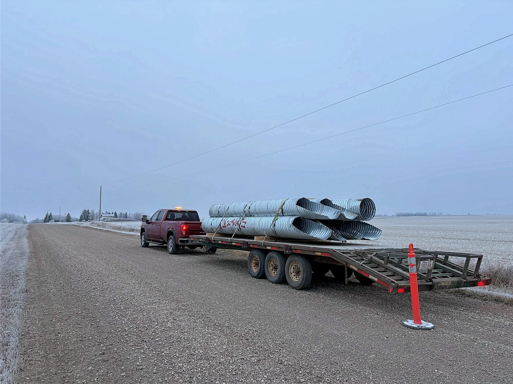 Red pickup truck parked on the side of a gravel road, carrying a trailer loaded with large metal pipes, with a cloudy sky and a flat, open rural landscape in the background.