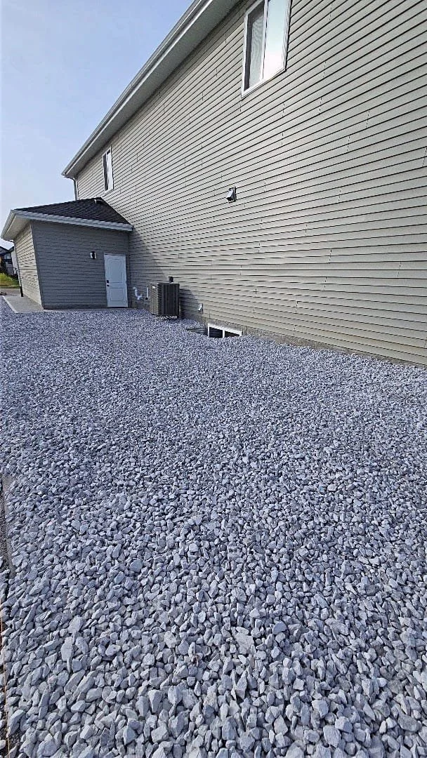 Side of a house with beige siding, a small gray shed, and a gravel yard with gray stones.