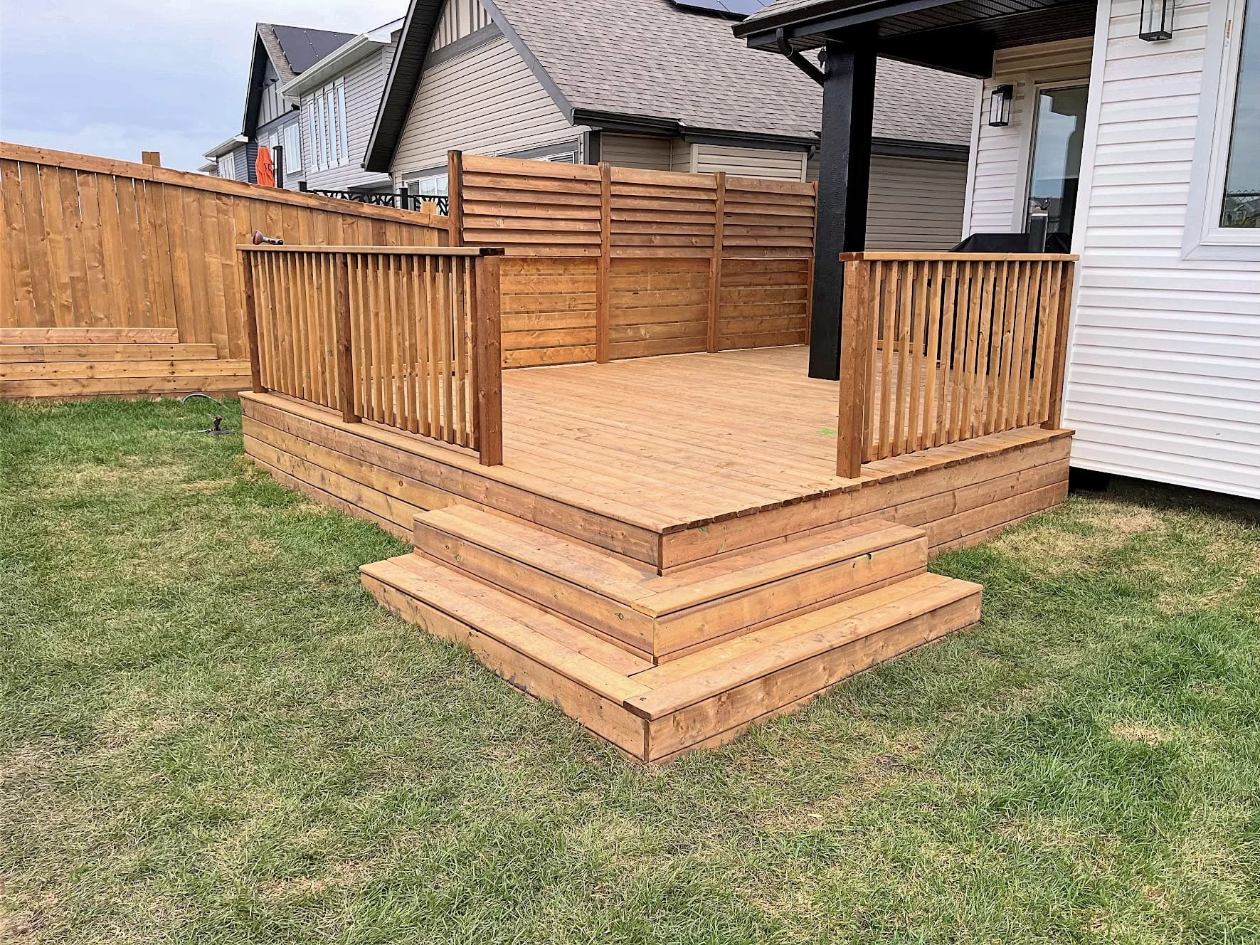 Newly built wooden deck with stairs and railings in a backyard, surrounded by grass and neighboring houses.