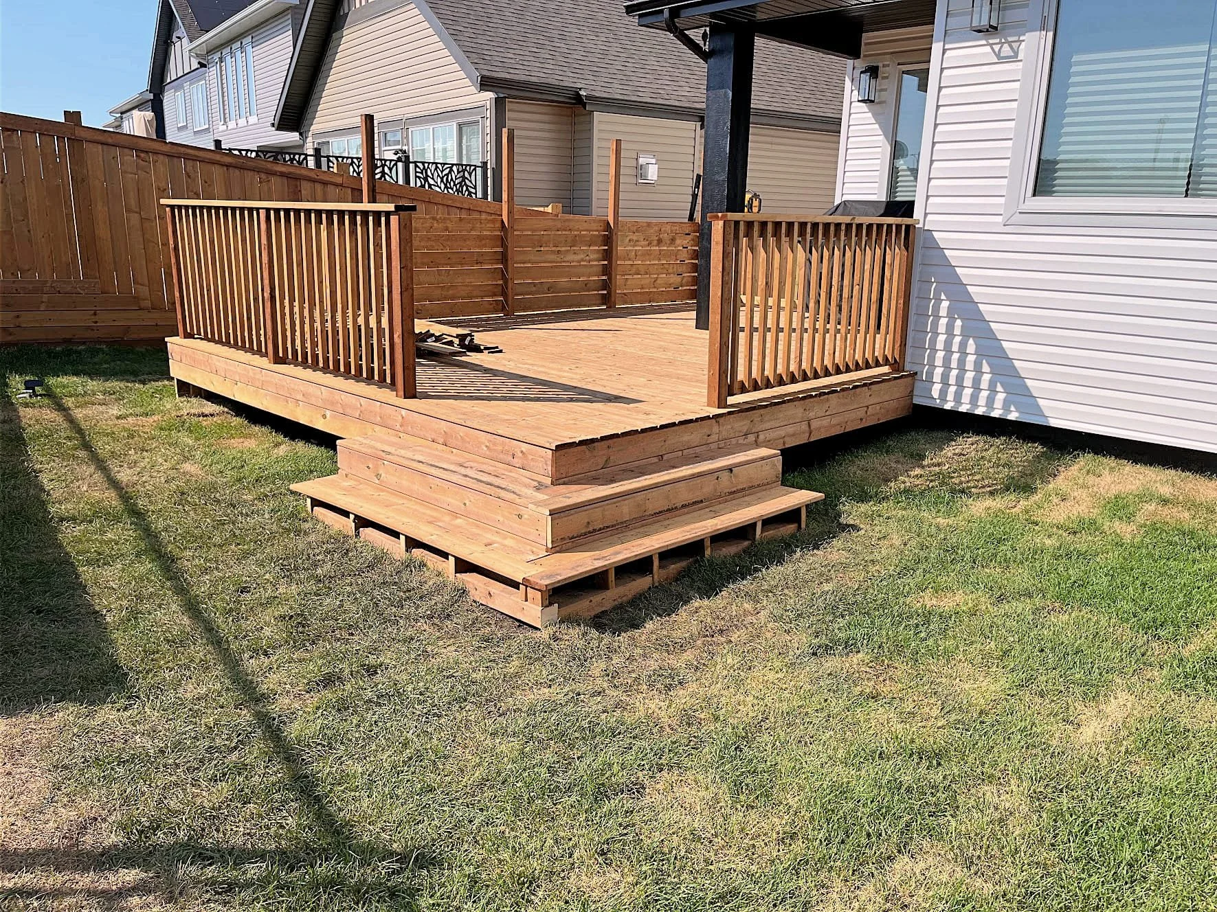 Newly built wooden deck with stairs and railings attached to a white house, surrounded by a grassy backyard.