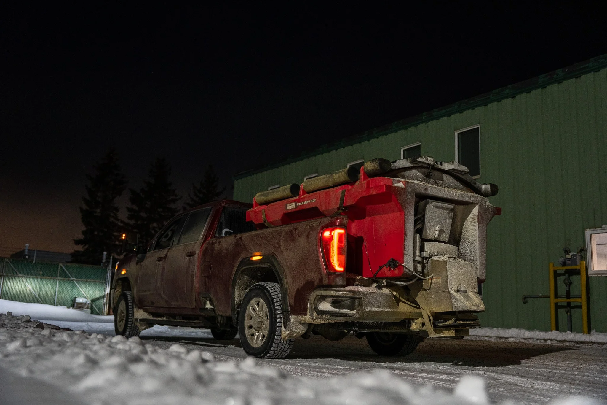 A yellow John Deere snowplow clearing snow at night with a dark sky and a building in the background.