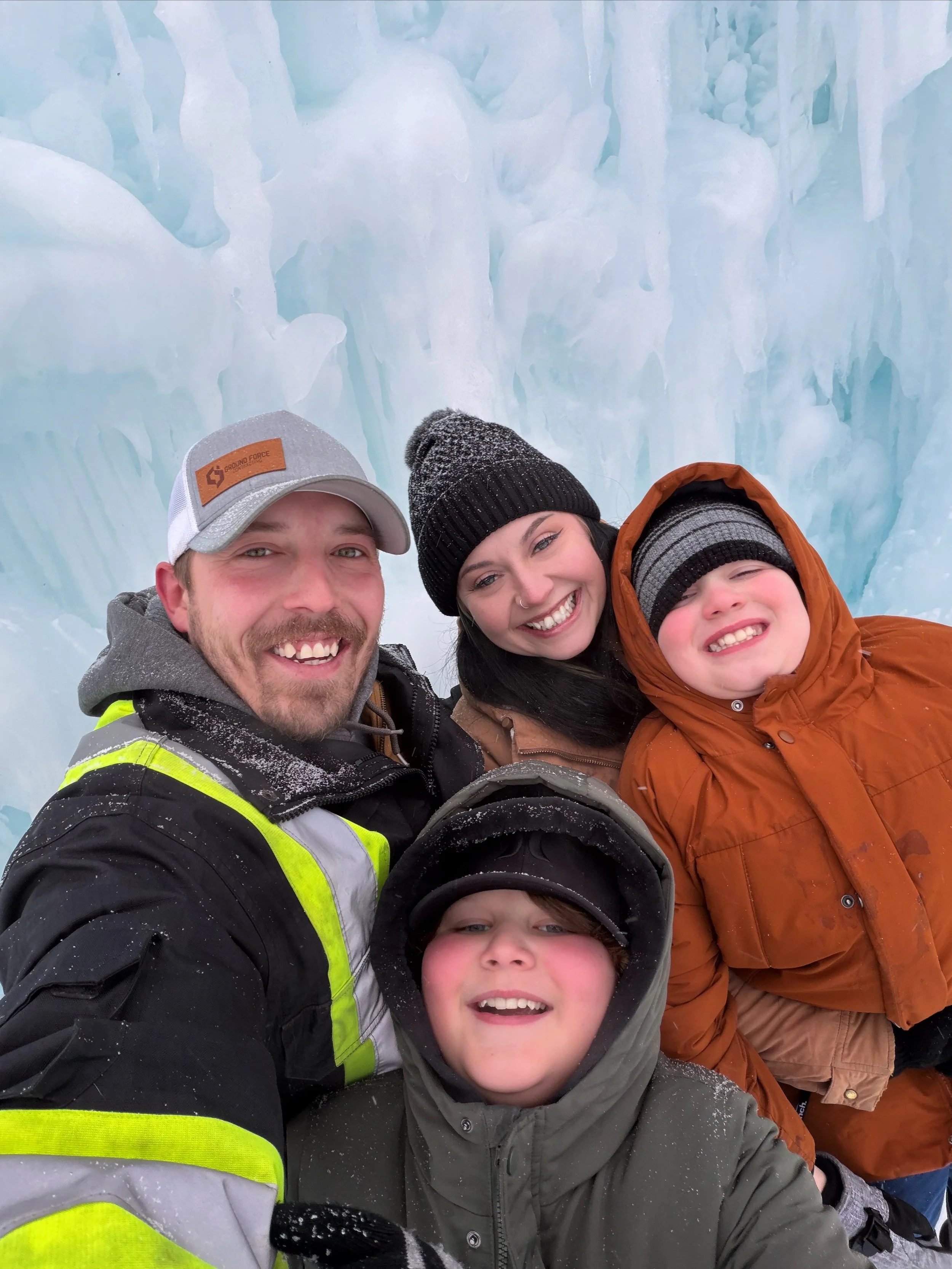 A group of four people smiling and taking a selfie in front of ice formations, dressed in winter clothing and hats.