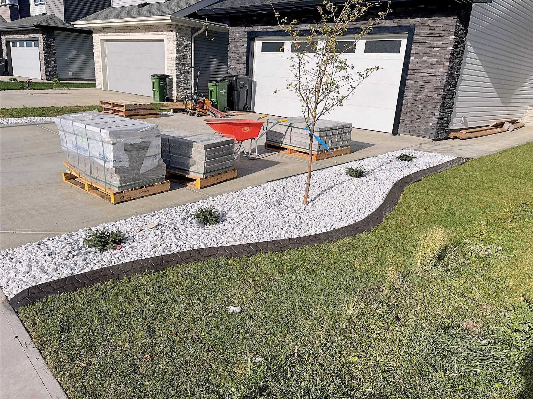 Front yard with white gravel landscaping, new small tree, and plants surrounded by dark edging. Construction materials and tools on driveway in front of a modern house with dark brick and beige siding, two white garage doors, and green trash bins.