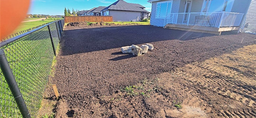 A backyard with freshly laid dark soil or compost, some small plants growing, a black chain-link fence on the left, and a white house with a porch on the right. There are a few bricks in the center of the yard.