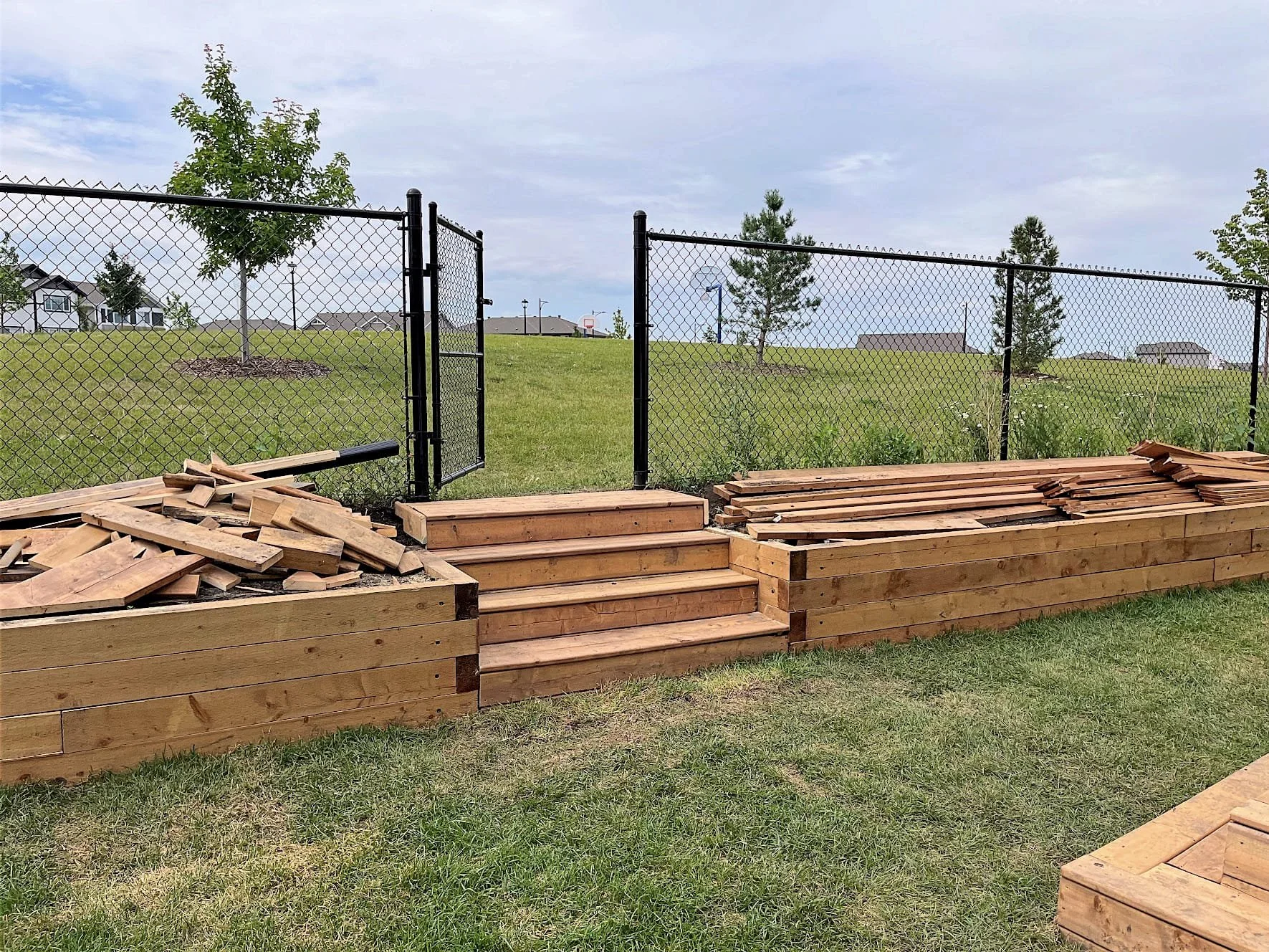 Wooden garden bed with steps leading up to a fenced grassy area with trees and houses in the background.