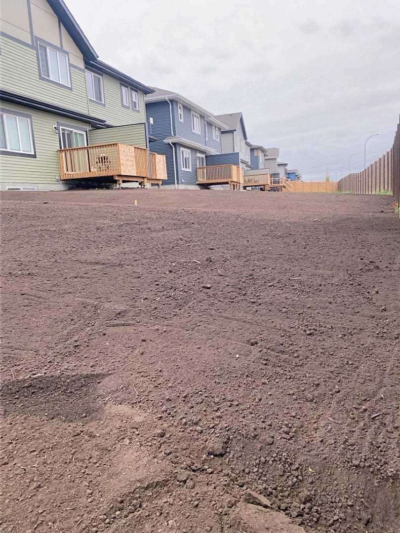 View of residential houses with small wooden decks, above a large area of freshly tilled or bare dirt, on a cloudy day.