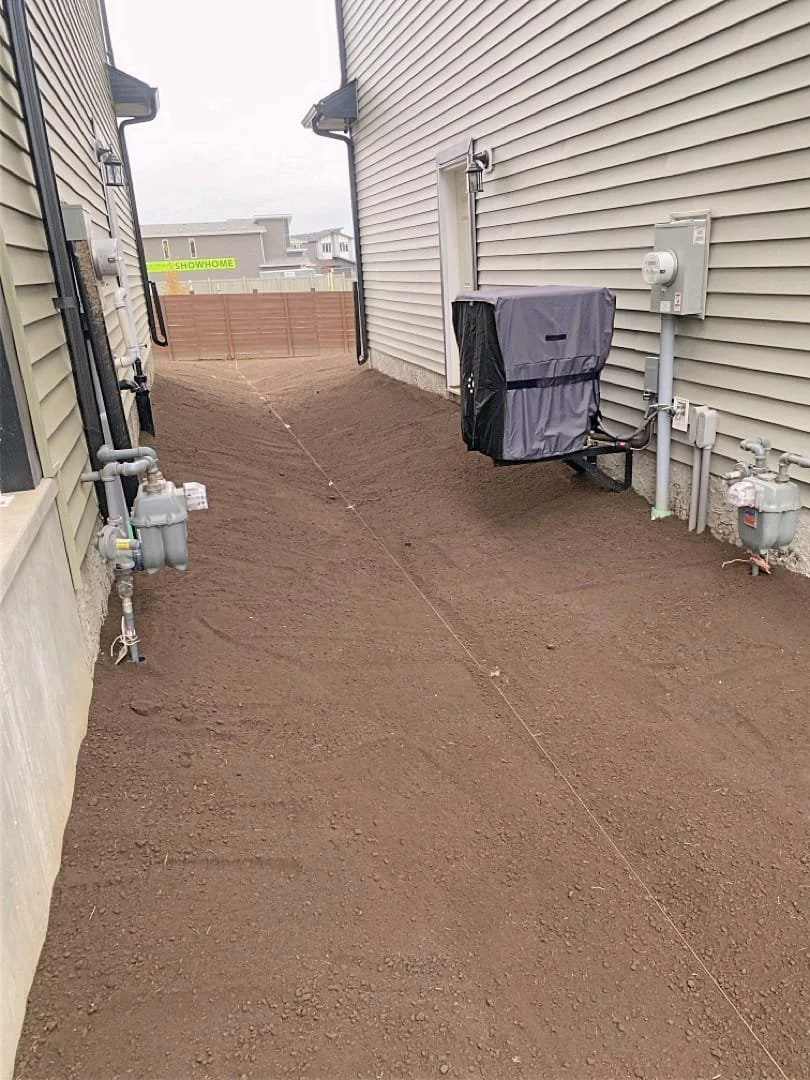 Backyard area with leveled dirt ground between two houses, utility meters and pipes on the right side, and a fence in the background with a 'SHOWHOME' sign.