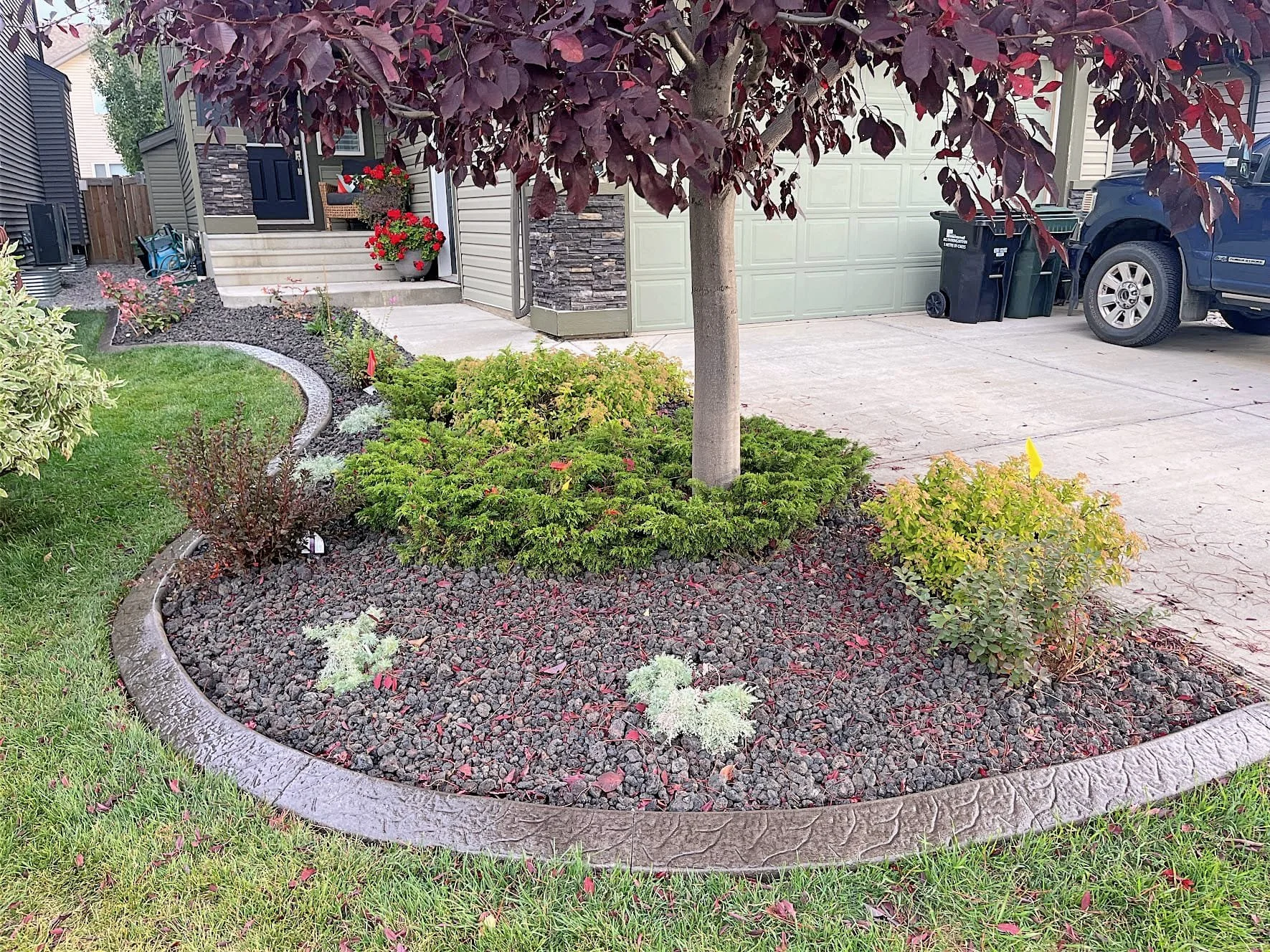 A landscaped front yard with a tree in the center, surrounded by various bushes and plants, with a curved stone border separating it from a grassy lawn. The house has a porch with potted red flowers, and a driveway with a black truck parked, along wi