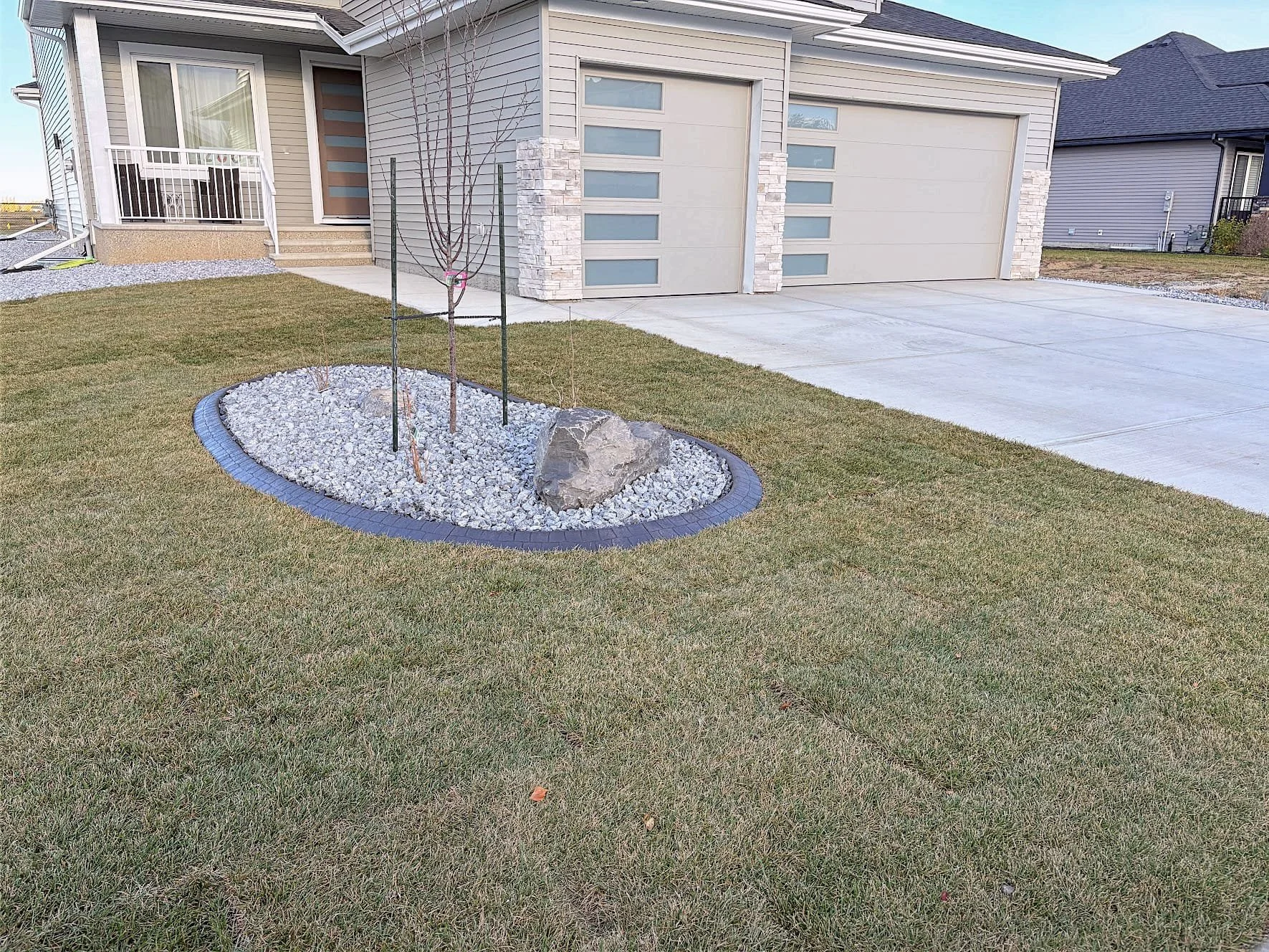 Front yard with a small landscaped garden bed with a young tree, rocks, and gravel, next to a modern house with a garage and a concrete driveway.