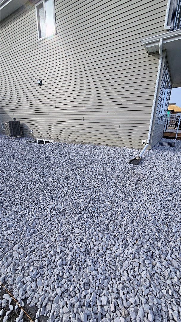 Side of a beige house with beige vinyl siding, a small window, and an air conditioning unit outside. The ground is covered with gravel, and there is a downspout attached to the house.