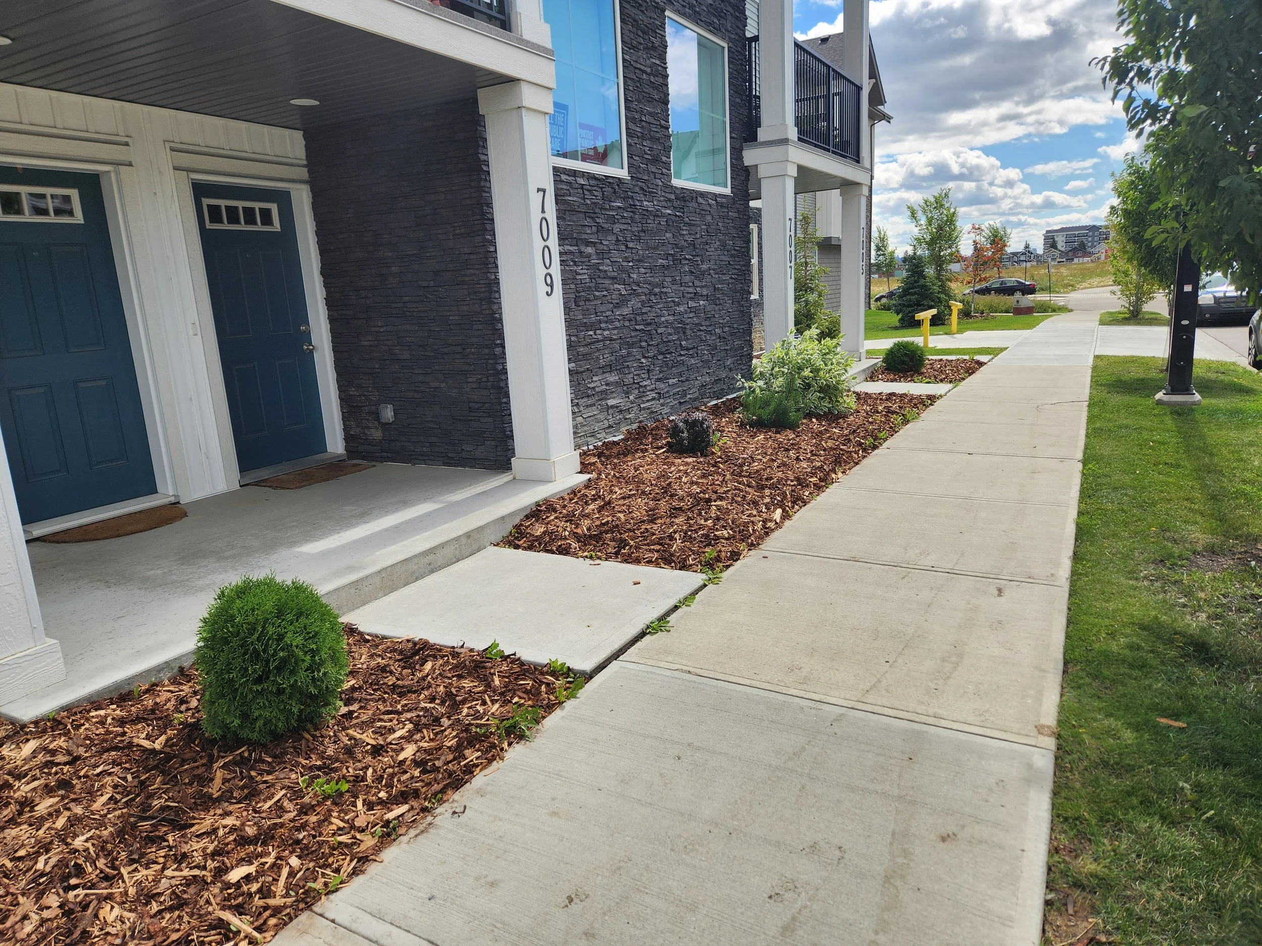 Sidewalk view of a modern apartment building with blue doors, white trim, and stone siding. Small landscaped garden beds with shrubs and mulch. Green grass, trees, parking lot, and cloudy sky in the background.