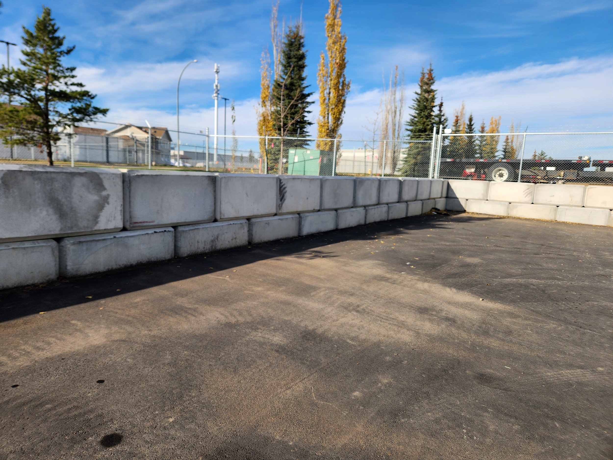 A construction site with a concrete block wall, asphalt ground, trees, and a blue sky with clouds.