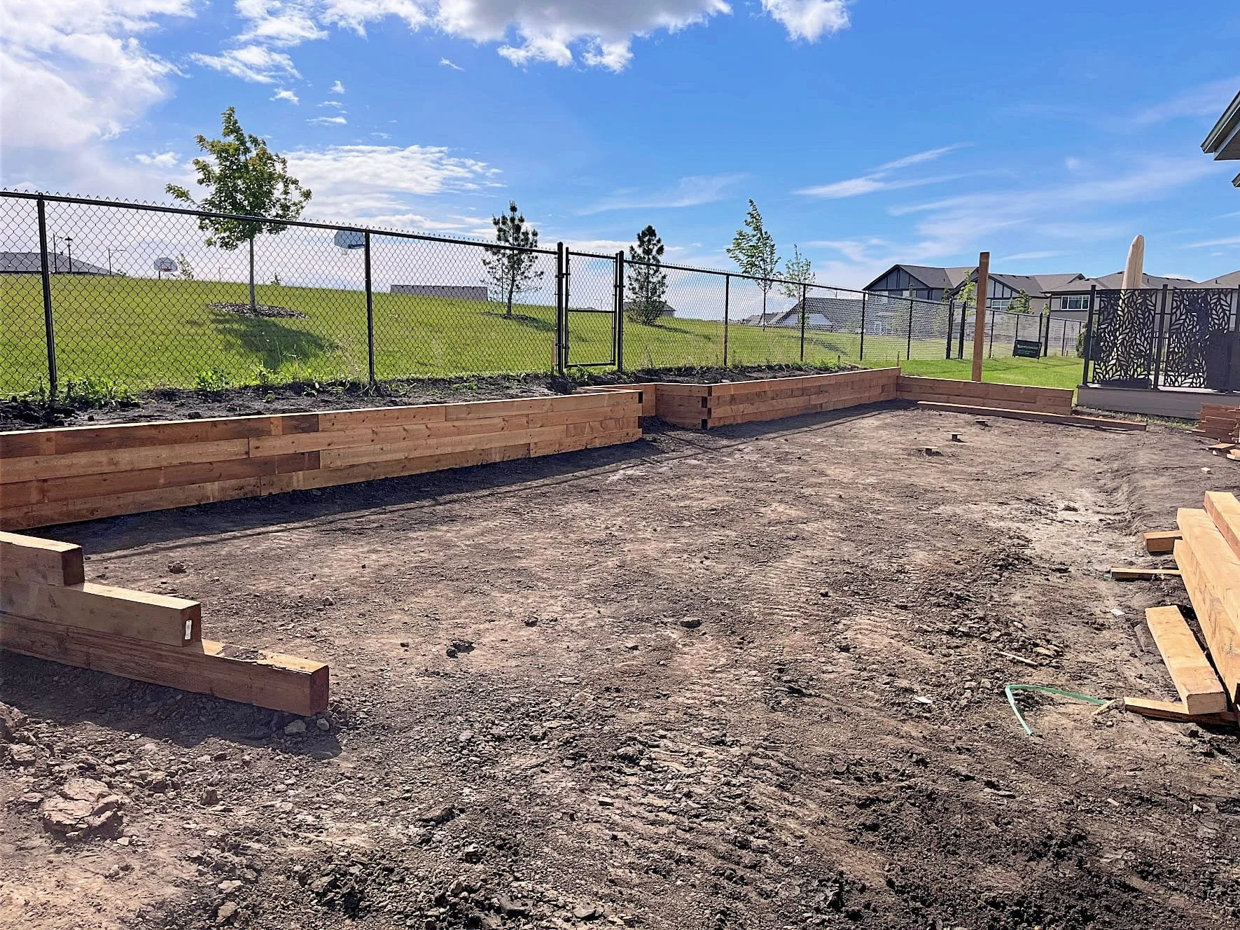 Construction site with wooden landscaping borders, dirt ground, and a black metal fence in the background, with a grassy hill and houses in distance under a blue sky with clouds.