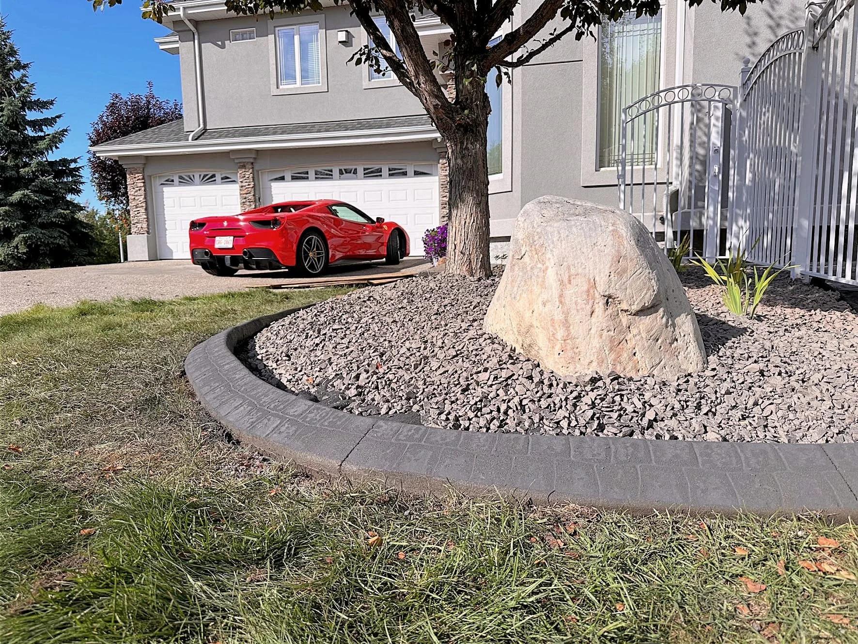 A red sports car parked in front of a gray house with a white garage door. The landscaped yard features a small tree, a large rock, and some plants, with a black paved border surrounding the gravel area.