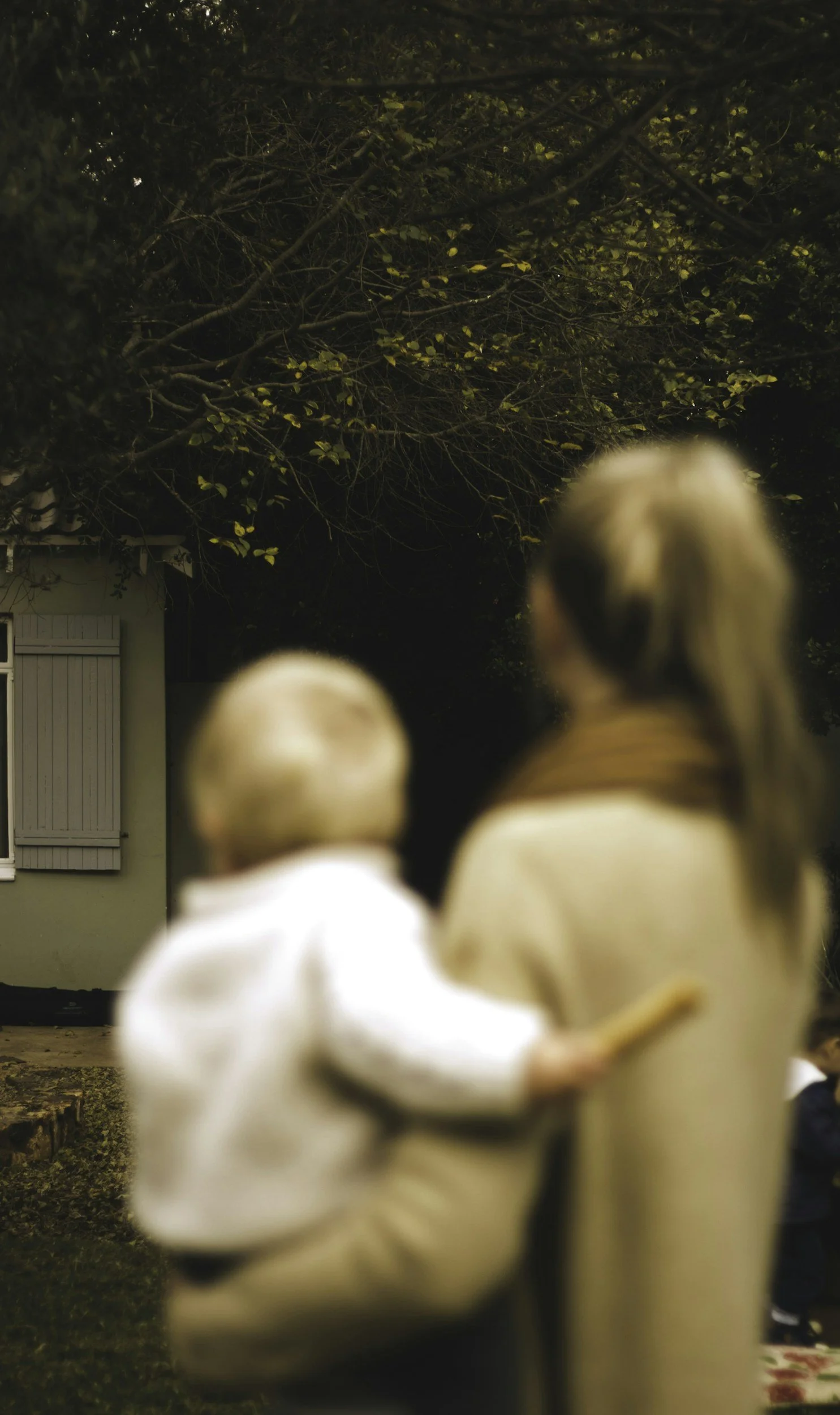 Two children, a girl wearing a martial arts uniform with a black belt and a boy in a white shirt, seen from behind, outdoors at night with a tree and a house in the background.