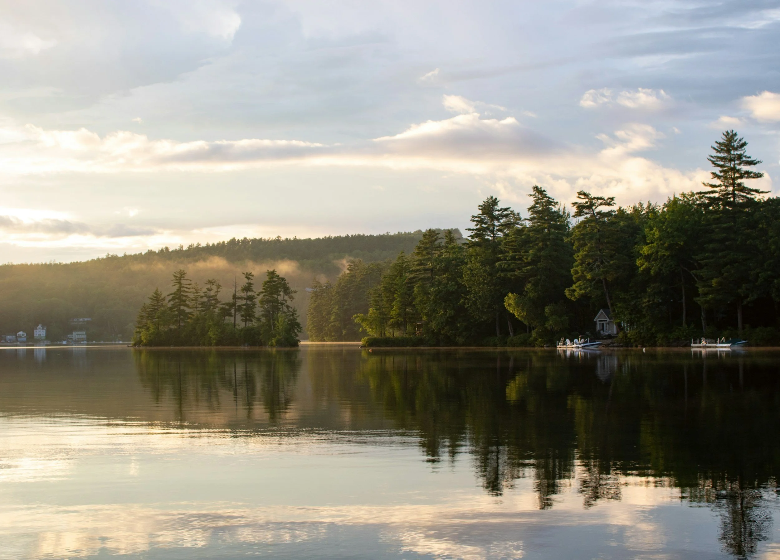Calm lake with reflection of trees and cloudy sky, houses and boats along the shore.