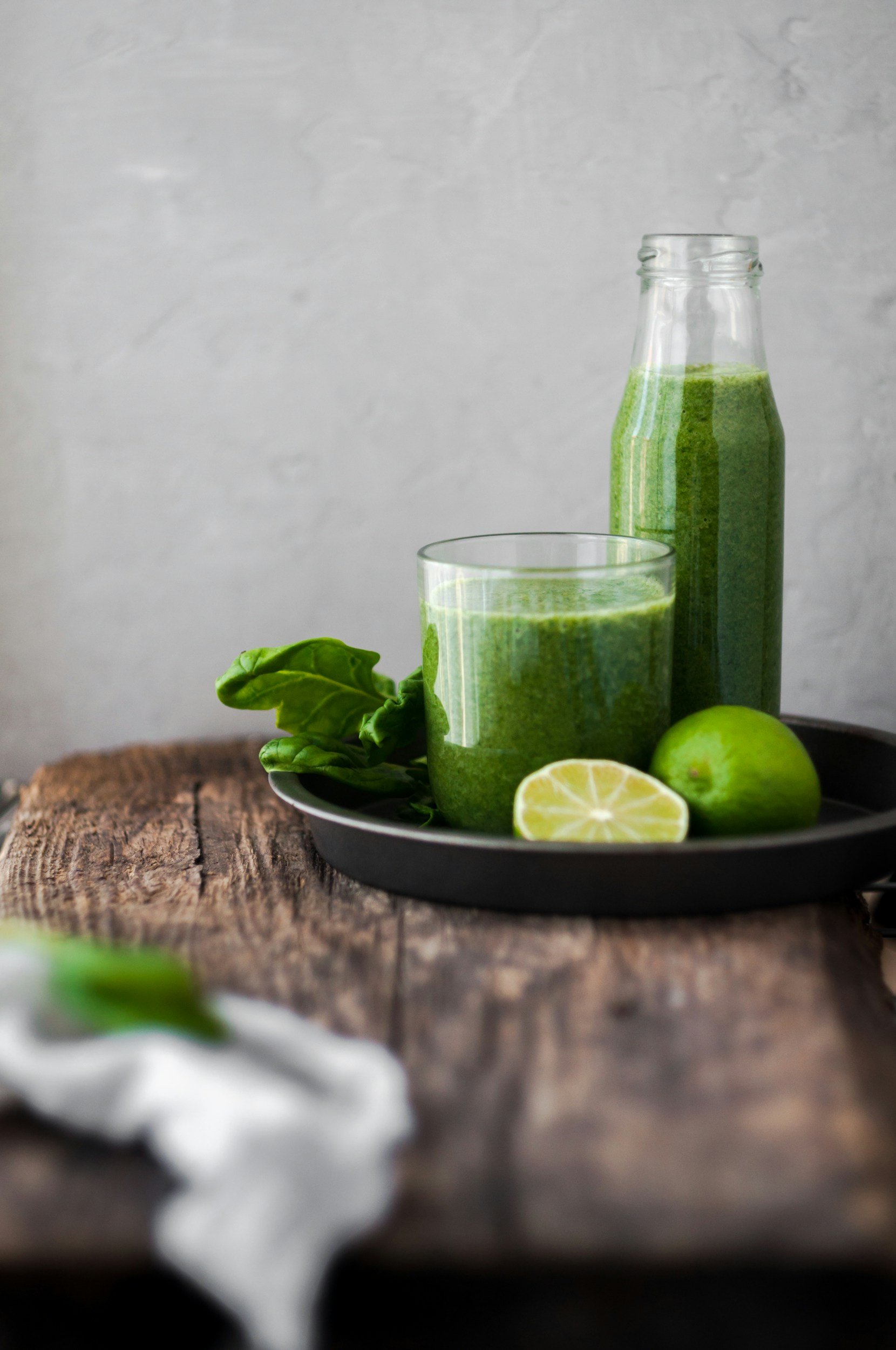 A glass jar filled with green smoothie, a glass of the same smoothie, fresh lime slices, a whole lime, basil leaves, and a dollop of white yogurt on a black plate on a rustic wooden surface with a gray textured background.
