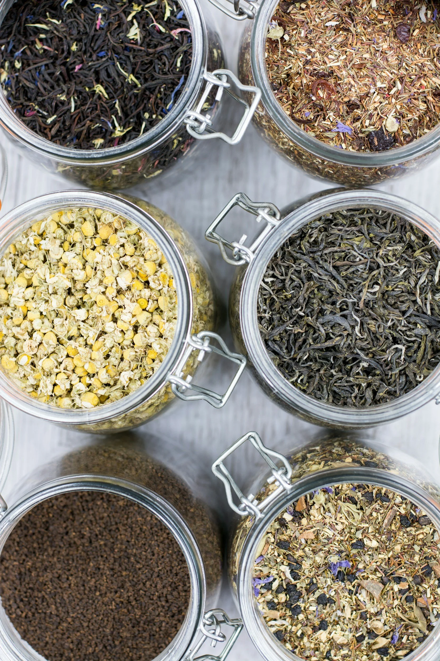 Six glass jars with metal clasps filled with various dried herbs, flowers, seeds, and loose tea leaves, arranged on a gray wooden surface.