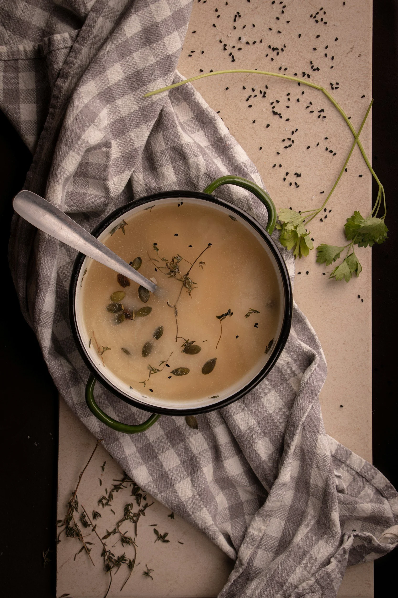 A bowl of herb-infused broth with herbs and seeds, surrounded by scattered herbs and seeds on a paper surface, with a checkered cloth for decoration.