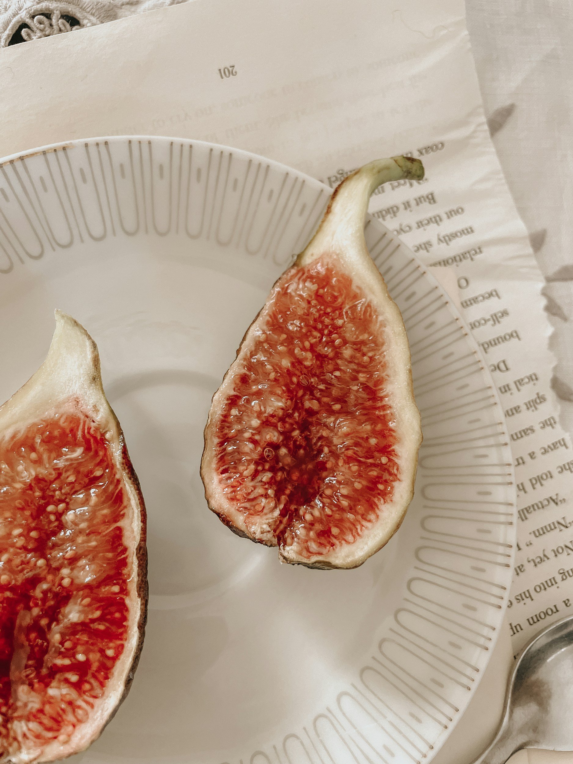 Close-up of two halved fresh figs on a white paper plate with printed text, part of a spoon is visible in the corner.