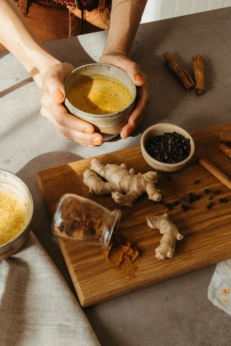 Hands holding a bowl of turmeric latte, surrounded by ginger, cinnamon sticks, black pepper, and turmeric on a cutting board.