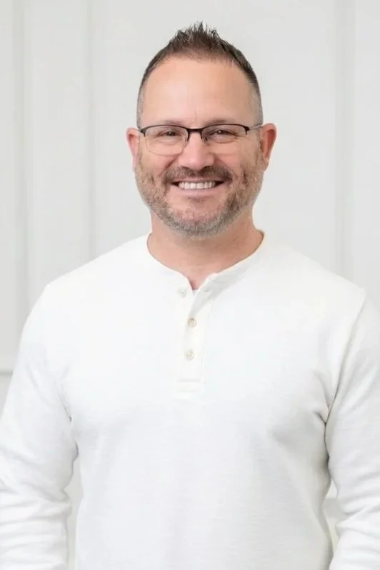 A smiling man, the owner of Emori Solutions with glasses and facial hair wearing a white long-sleeve shirt in front of a light background.