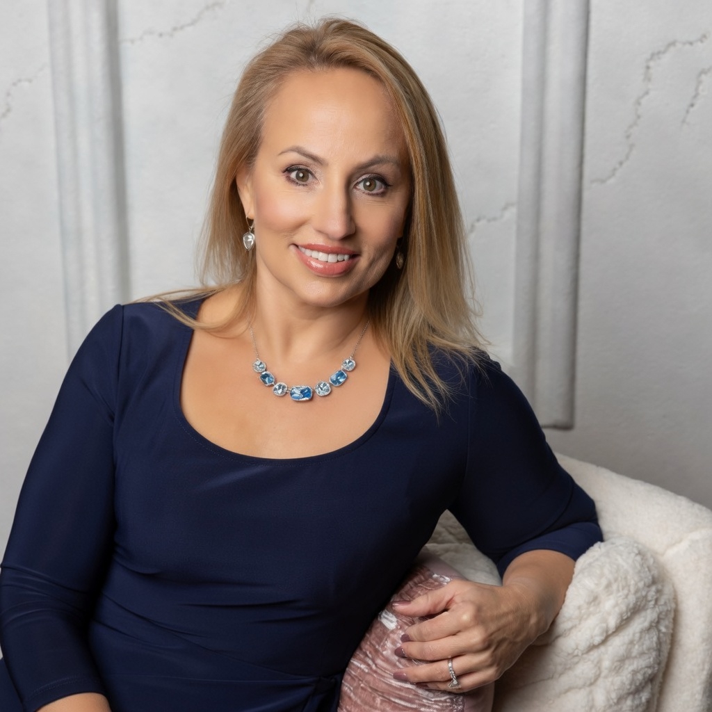 A woman, the owner of Emori Solutions, with blonde hair smiling, wearing a navy blue dress, necklace, earrings, and a ring, sitting on a white textured chair in front of a neutral wall.