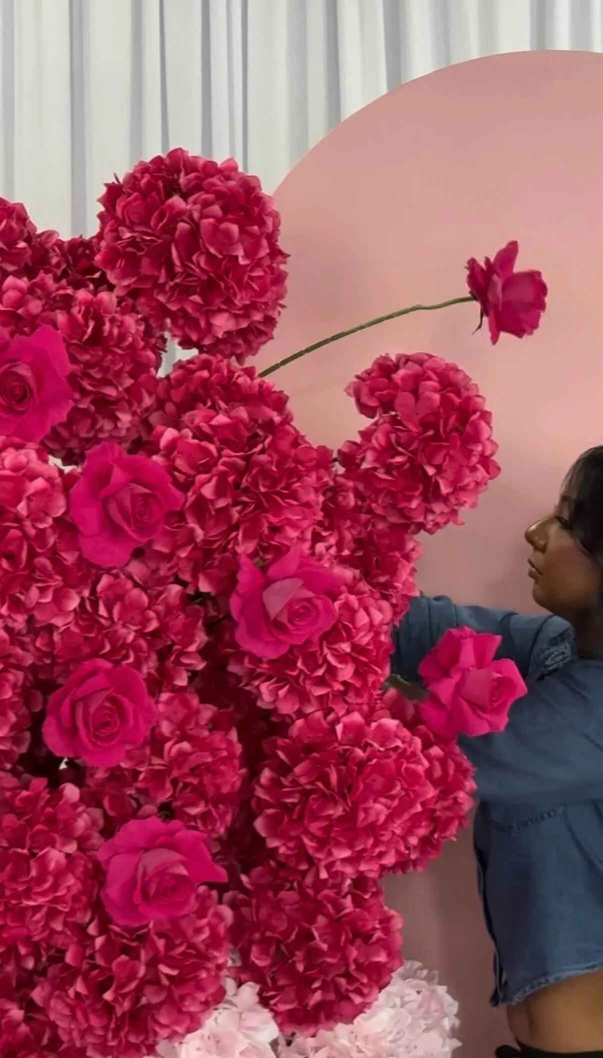 A woman with curly dark hair and glasses arranging pink and red flowers, including roses and hydrangeas, in front of a light pink backdrop.