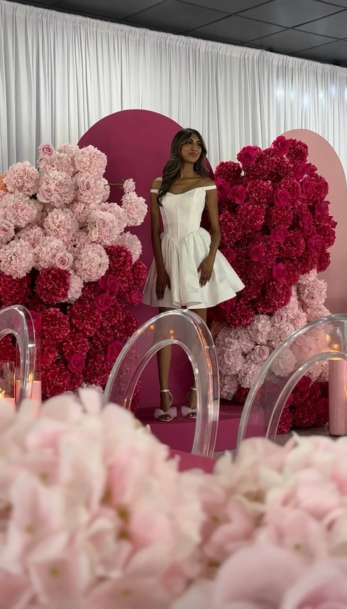 A woman in a white dress standing in front of a floral backdrop with pink, red, and light pink flowers and pink heart-shaped decor at an event.