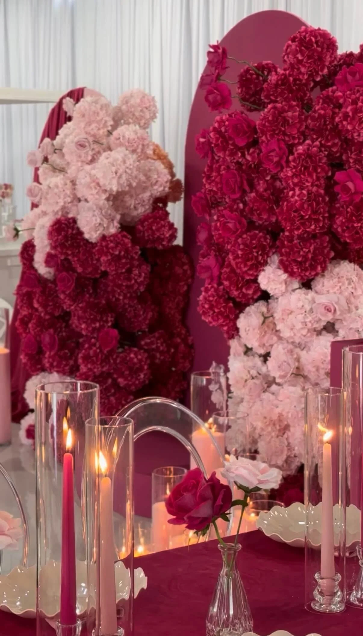Decorative table with pink and red flowers, candles, and a pink backdrop at an event or celebration.