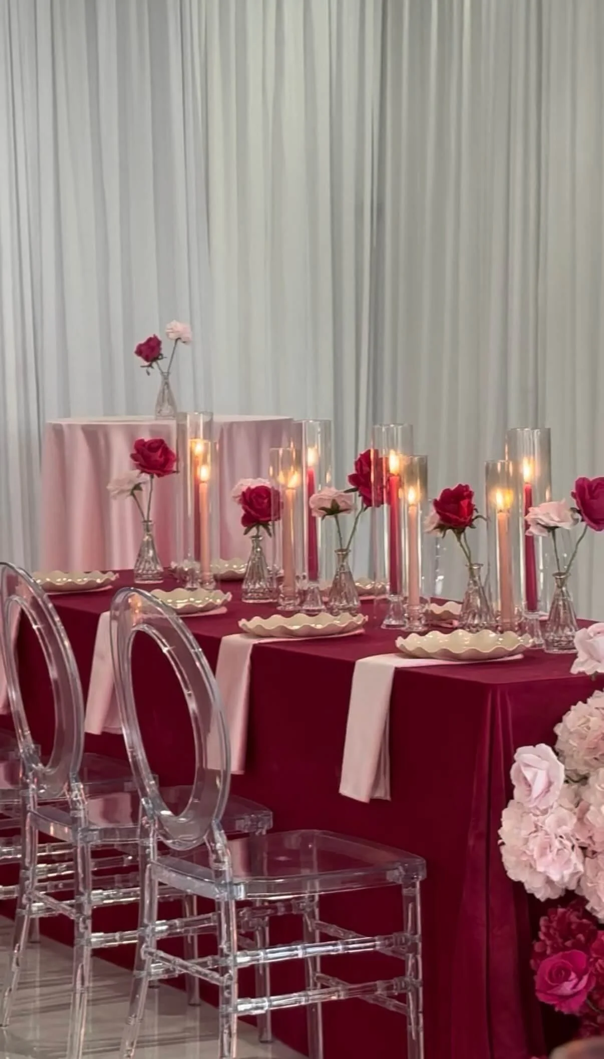 Decorated table with pink and red roses in glass vases, tall candles, and a pink and burgundy tablecloth, set for an event with a white curtain backdrop.