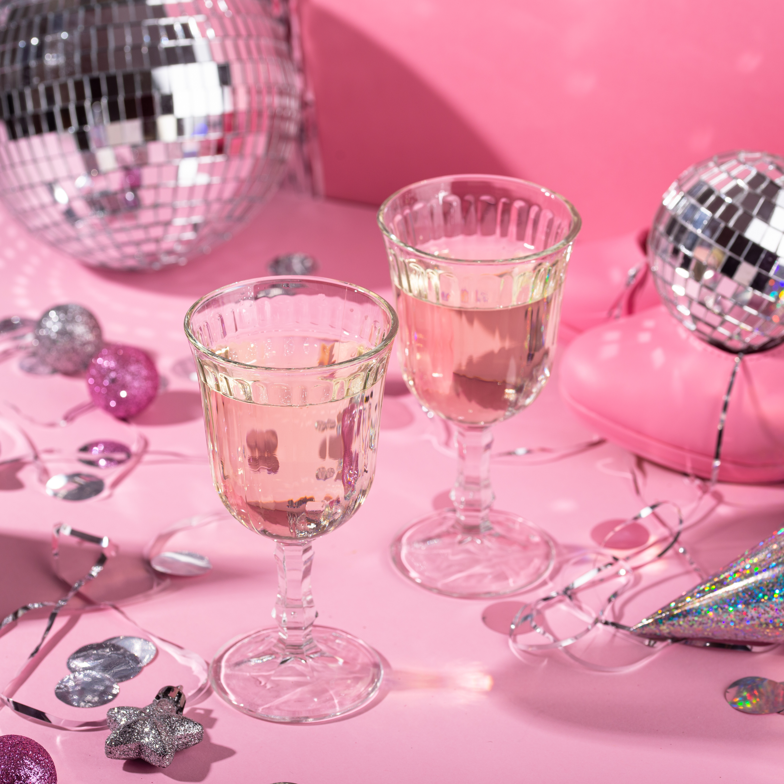 Two glasses of pink wine surrounded by pink and silver party decorations, including disco balls, glittery ornaments, and tinsel, on a pink surface.
