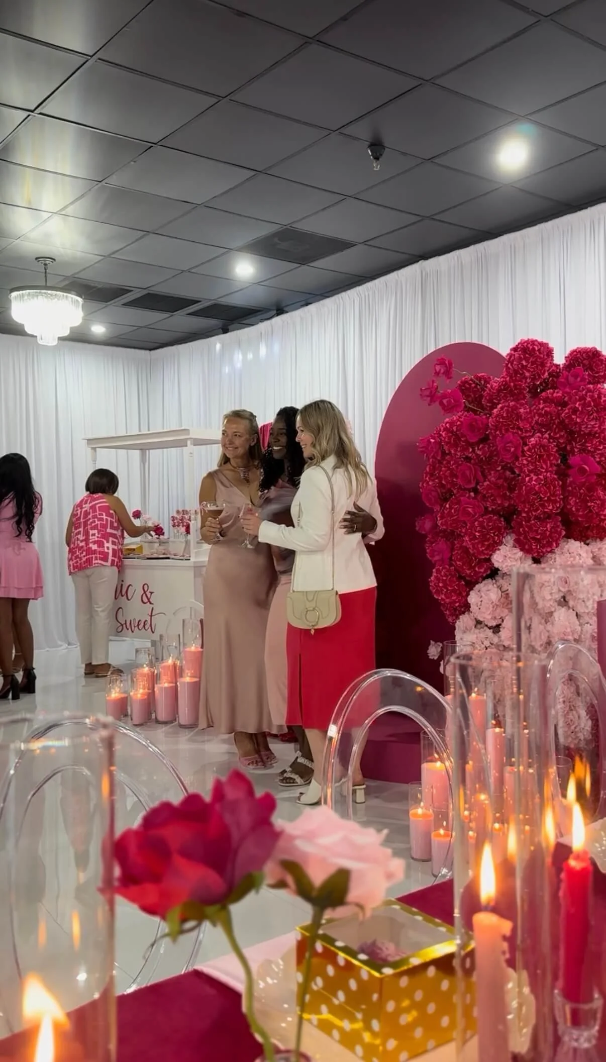 Women celebrating at a party decorated with pink flowers, candles, and a heart backdrop.