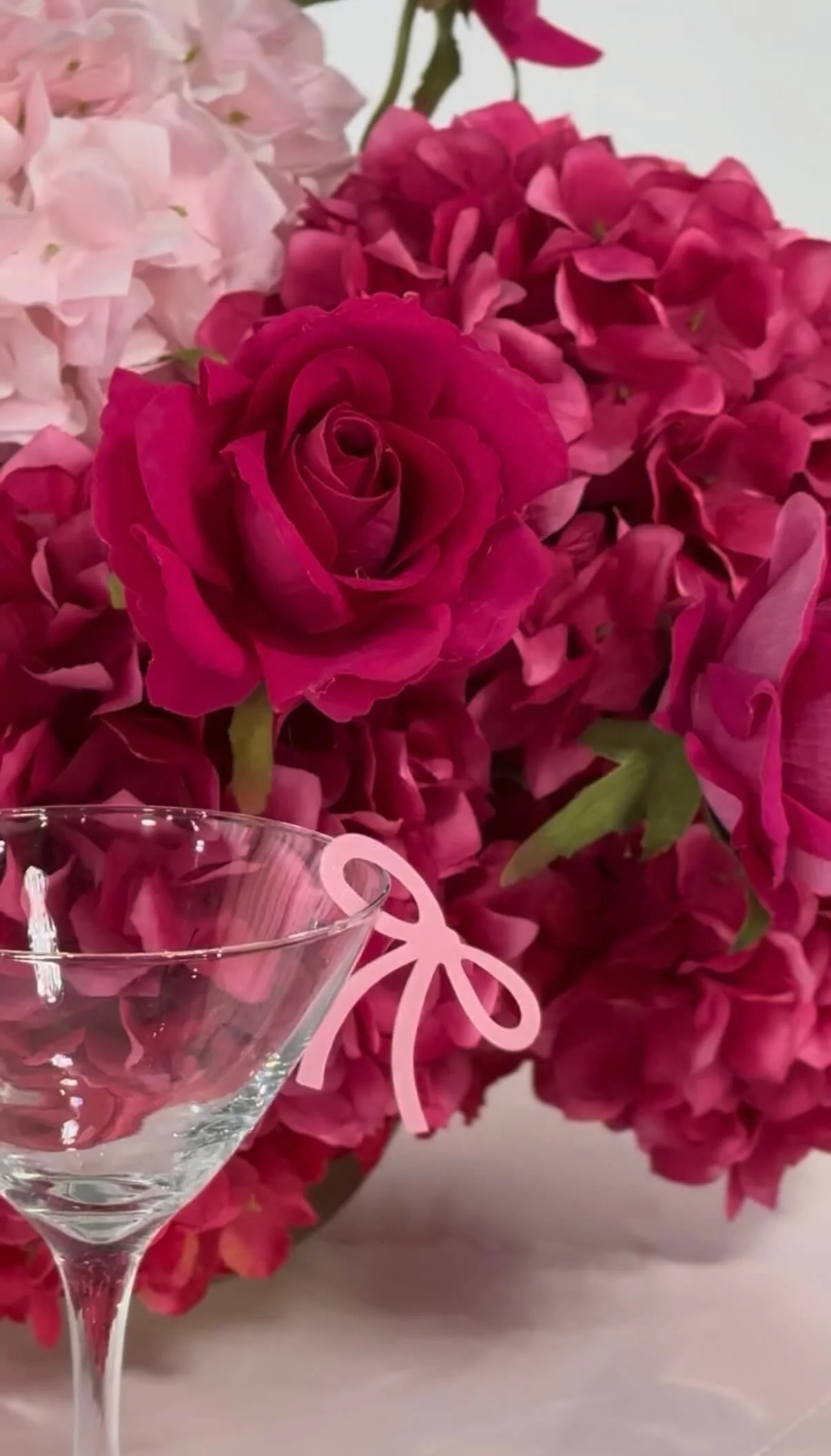 A close-up of pink and fuchsia hydrangea flowers with a clear martini glass in the foreground that has a pink ribbon decoration.