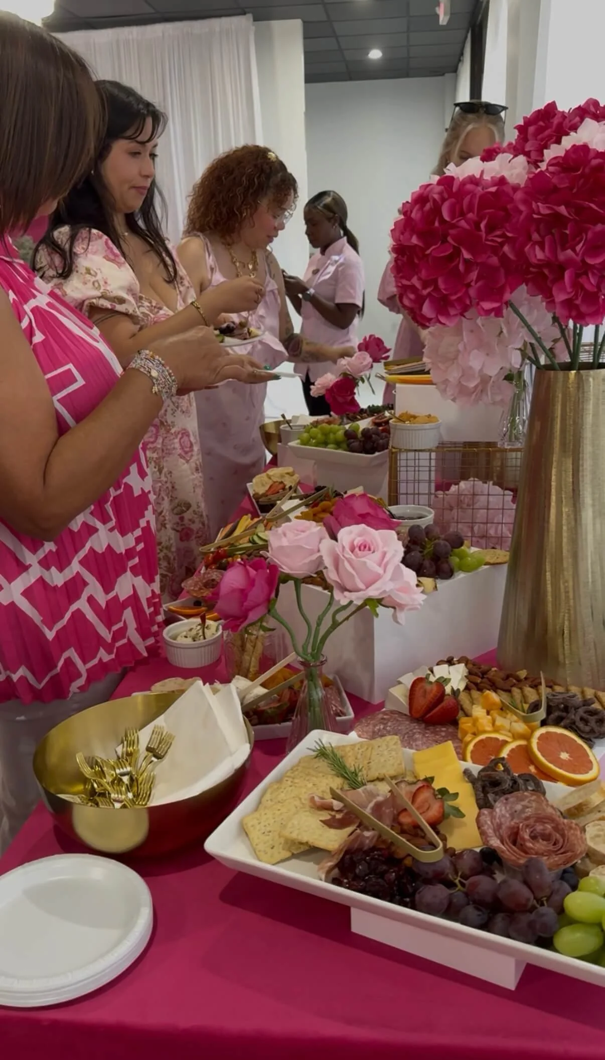 People serving themselves food at a pink-themed buffet table decorated with pink and white flowers.