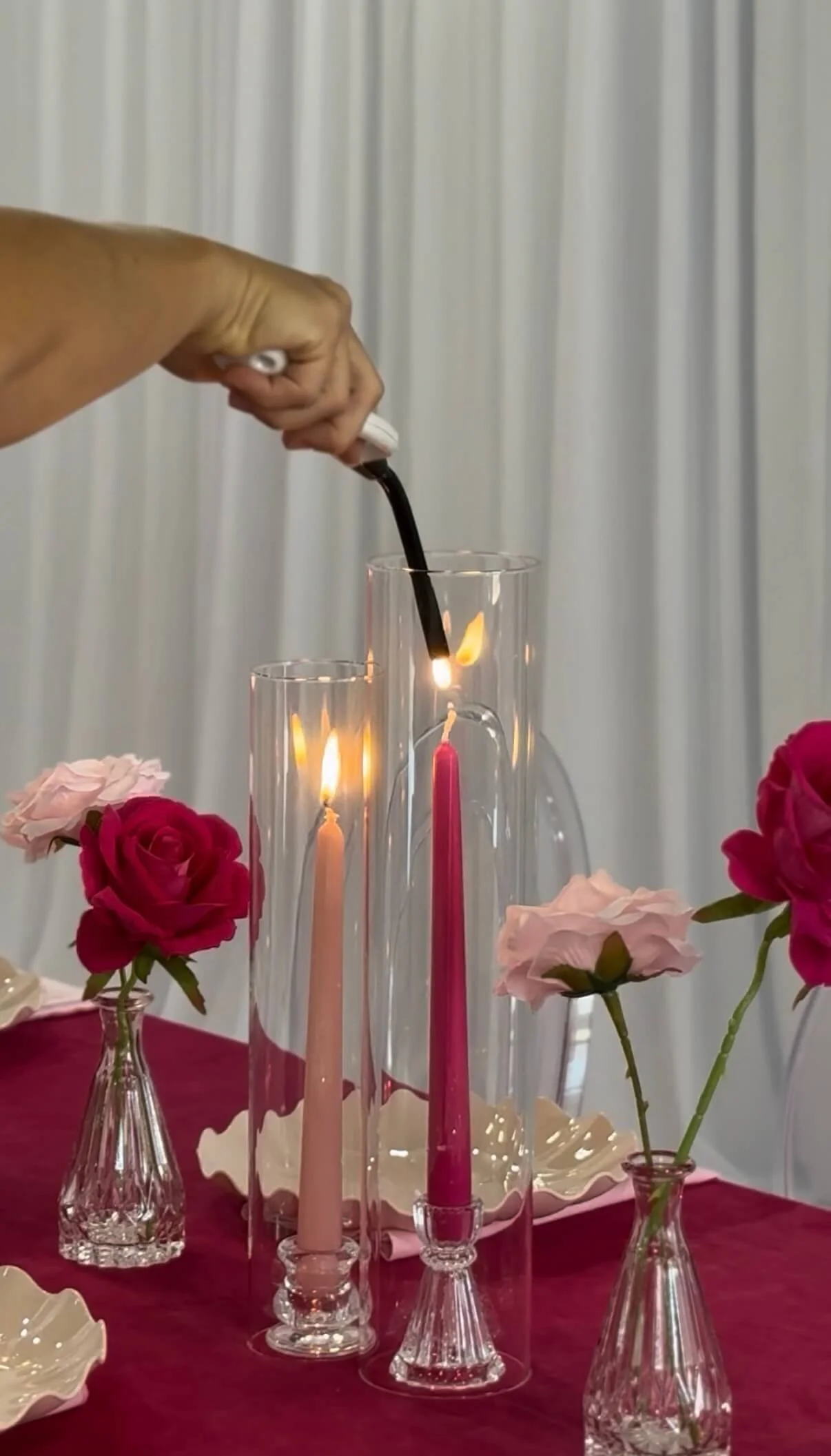 A person lighting a candle inside a glass hurricane on a table decorated with pink and red roses, and pink candles in glass candle holders, with a white curtain in the background.