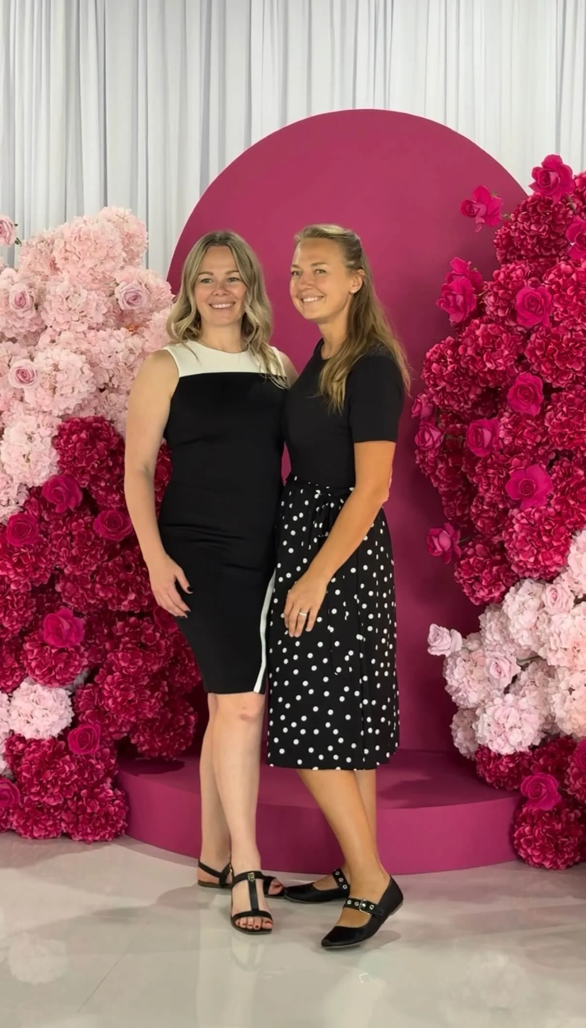 Two women standing close together in front of a pink floral backdrop with a pink circular display. Both women are smiling and wearing black shoes.