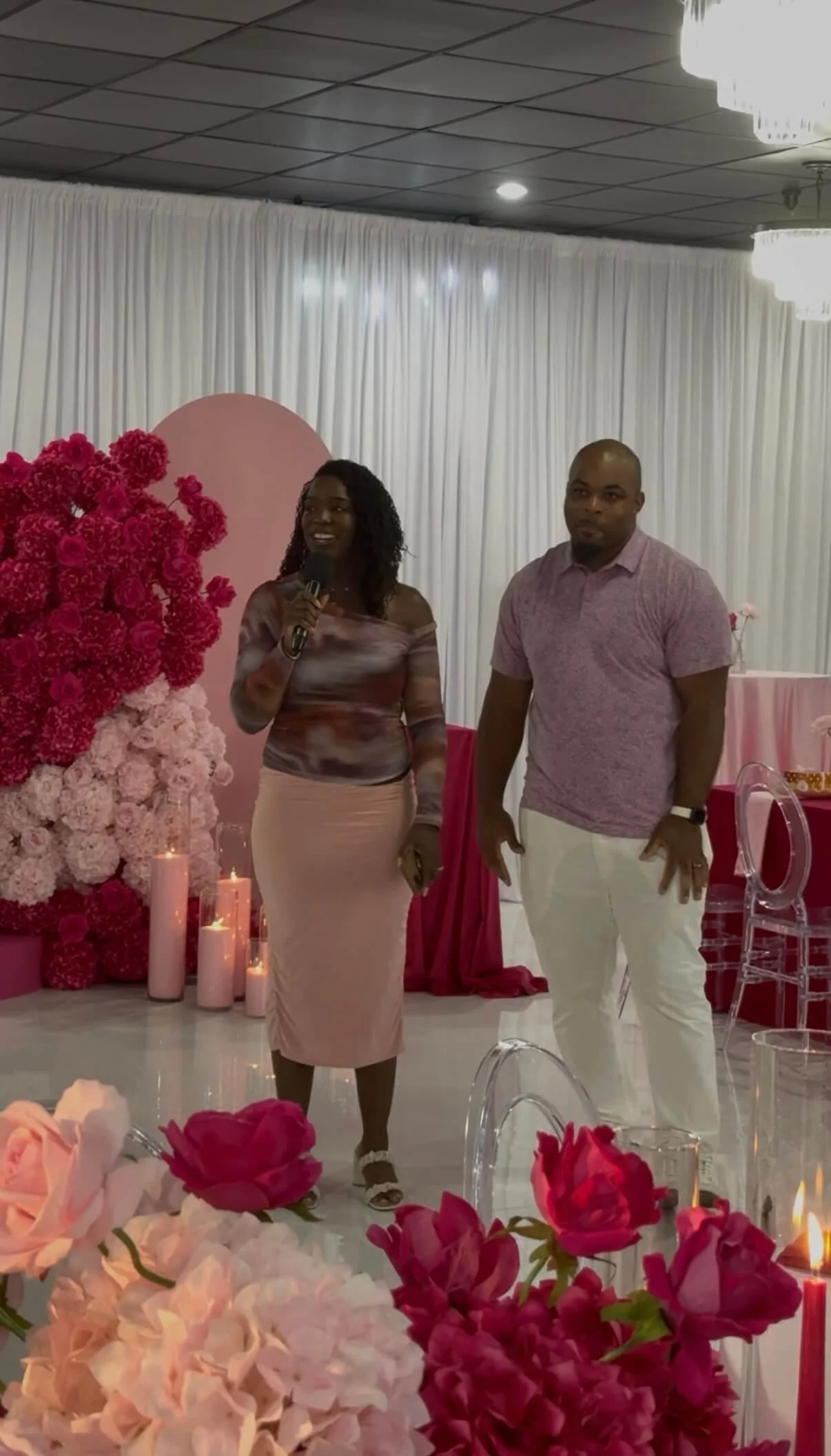 A woman holding a microphone and speaking at a decorated event with pink flowers, candles, and a pink backdrop, standing next to a man in a pink shirt and white pants.