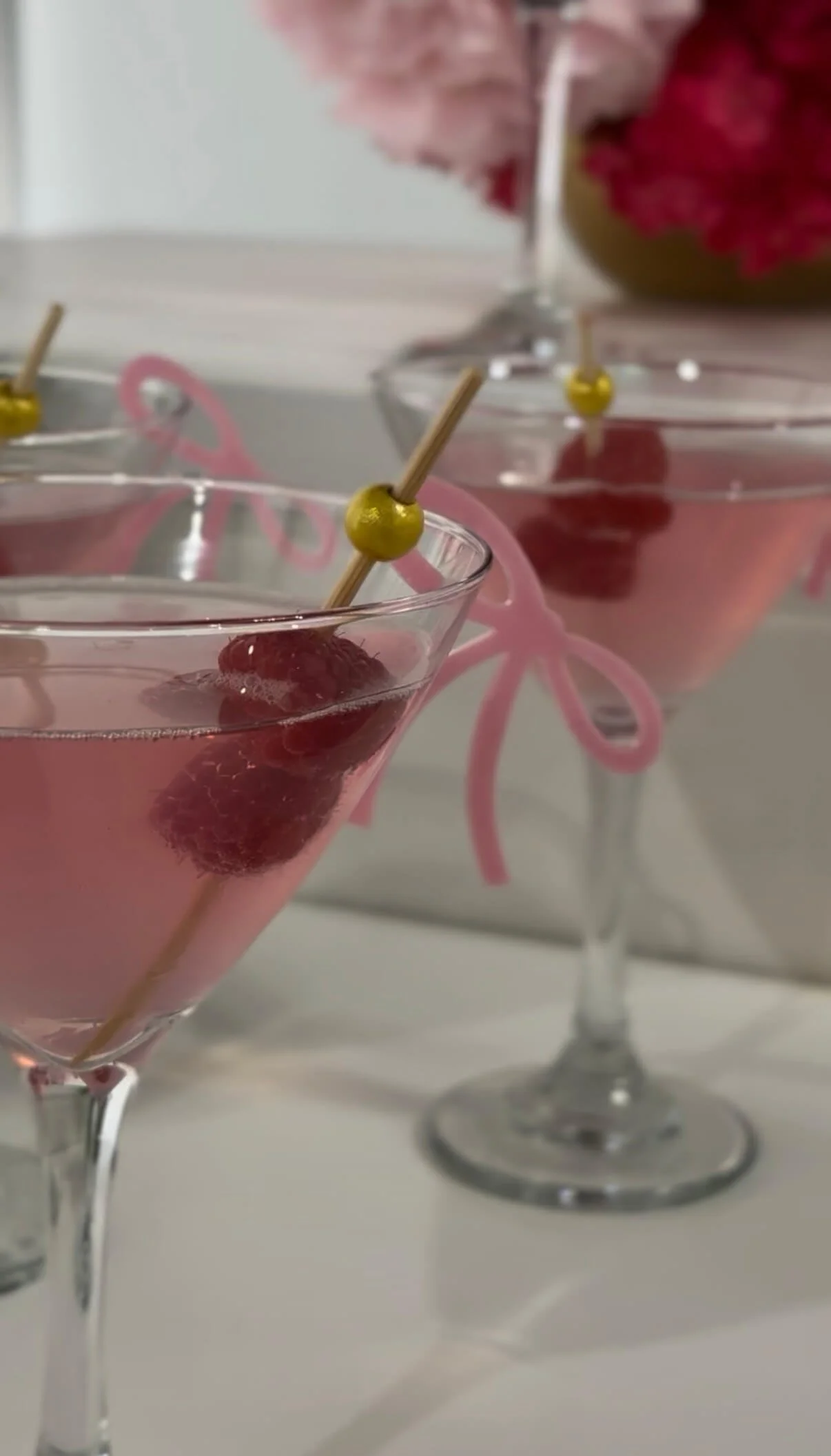 Three pink cocktails garnished with raspberries and topped with green olives on a skewer, served in crystal martini glasses.