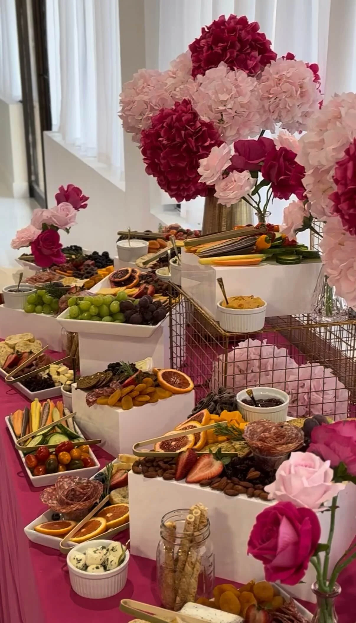 A table decorated with pink and red paper flowers holds a variety of foods, including fruits, vegetables, cheeses, and charcuterie, with a backdrop of white curtains.