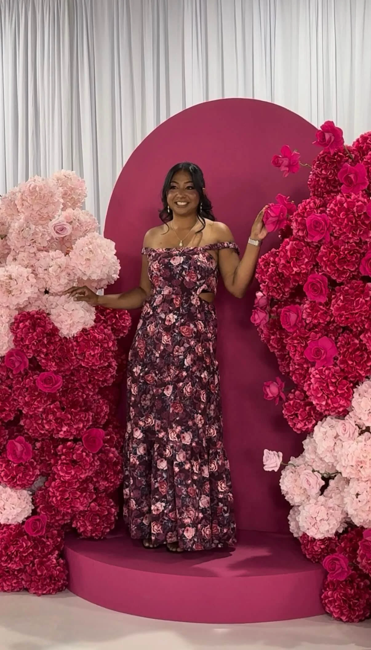 Woman in a floral off-shoulder dress standing among pink and red floral arrangements with a pink backdrop.