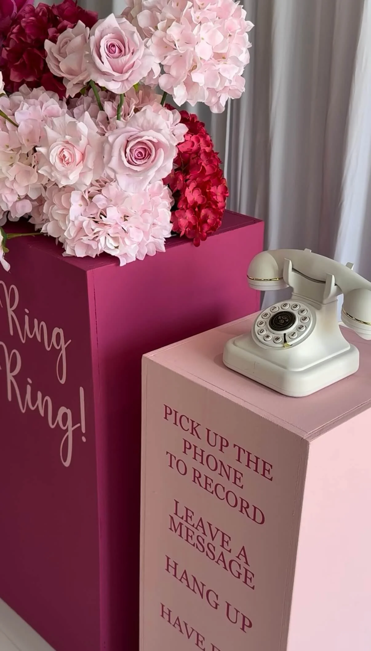 A vintage white rotary telephone on a pink box with red text, beside a larger pink box with pink and red hydrangea and rose flowers.